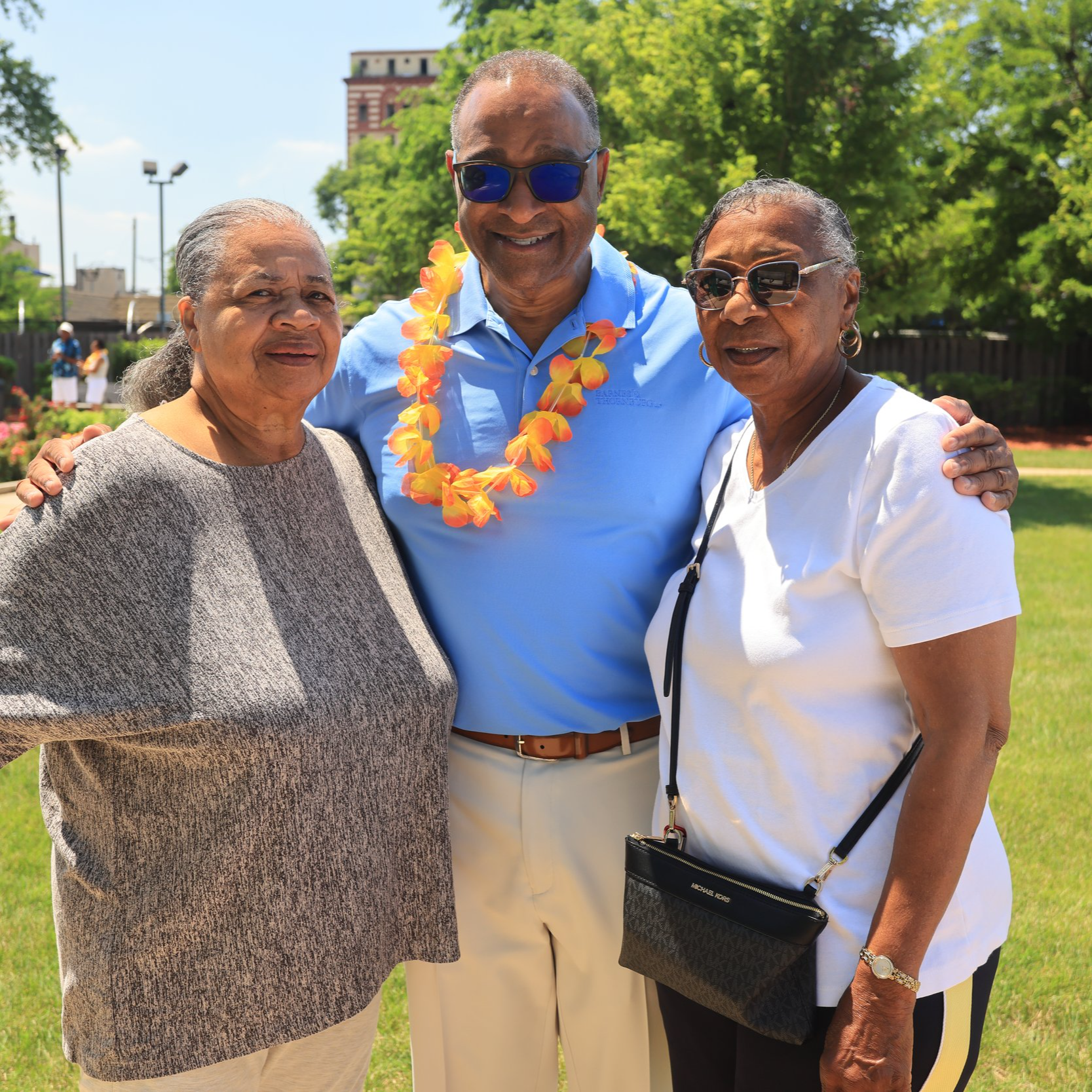Richard Boykin and two women, smiling and posing closely together outdoors on a sunny day. The man in the middle is wearing sunglasses, a blue shirt, and a colorful lei, with his arms around the women.