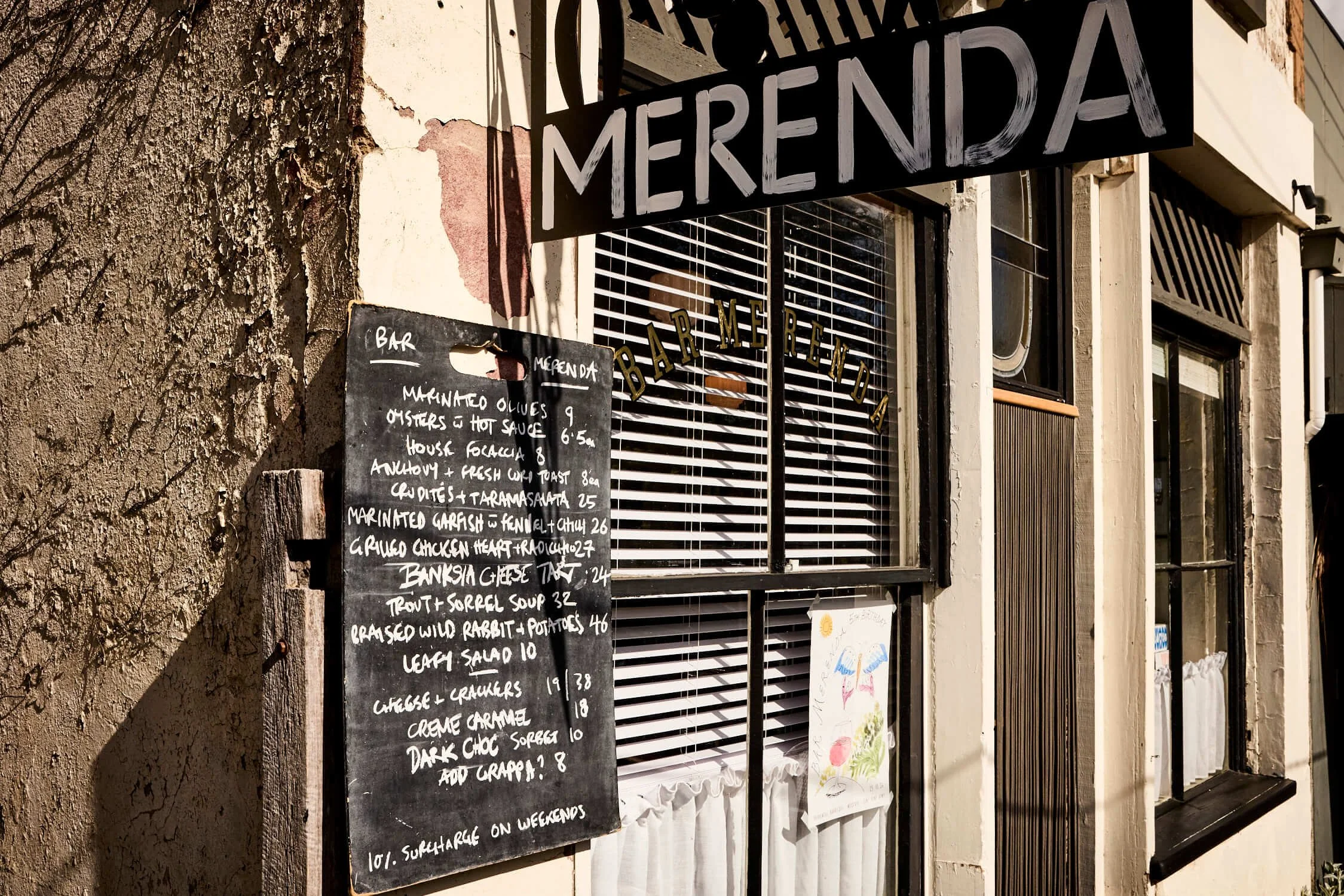 Exterior of Bar Merenda restaurant in Daylesford with a MERENDA sign, chalkboard menu, and striped window blinds.