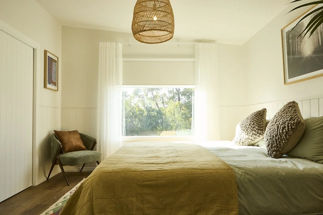 Bright cozy bedroom with a neatly made bed, textured pillows, sheer curtains, woven pendant light, armchair, and sunny window