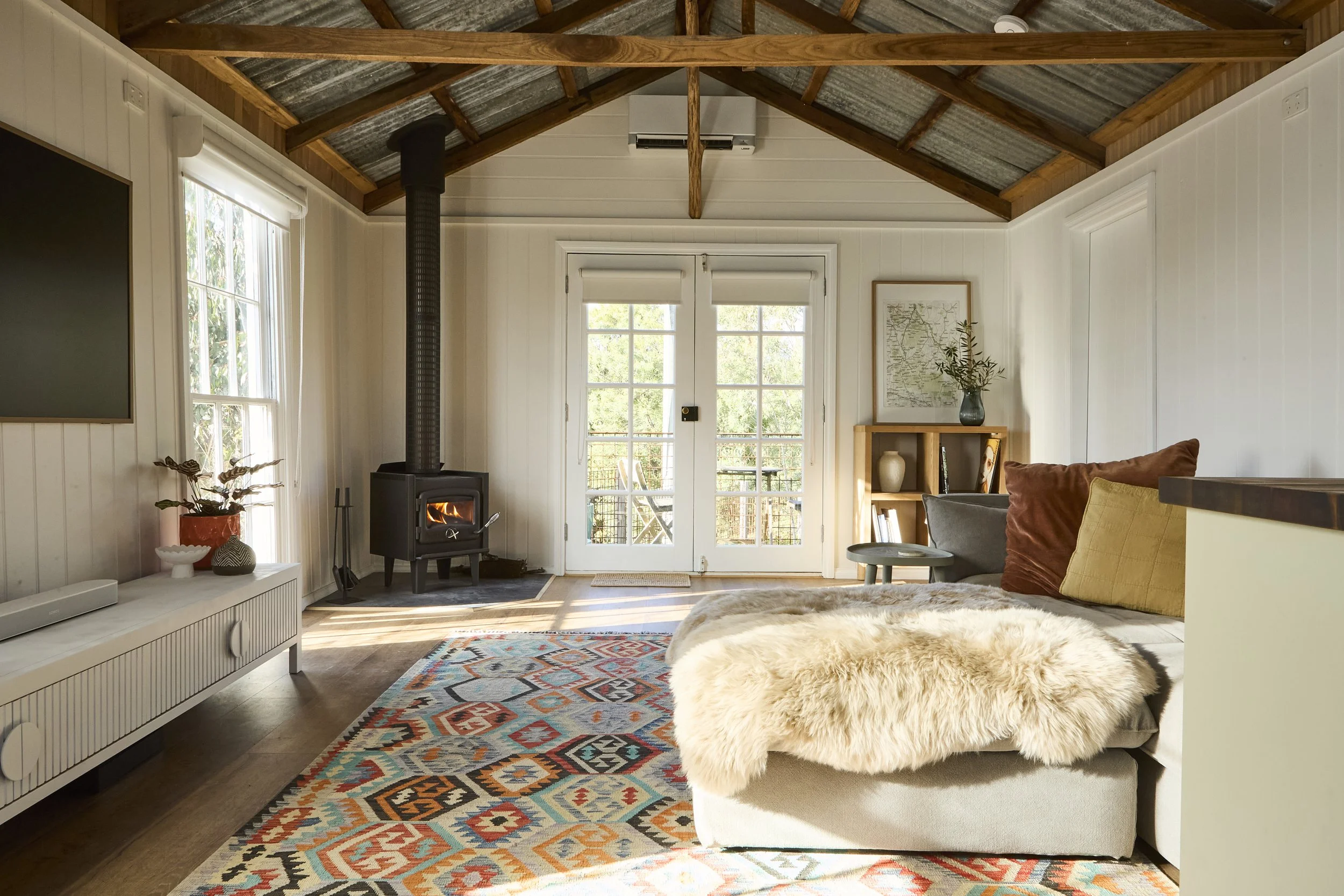 Bright rustic living room with a wood-burning stove, exposed timber beams, French doors, colorful patterned rug, and a gray sectional sofa