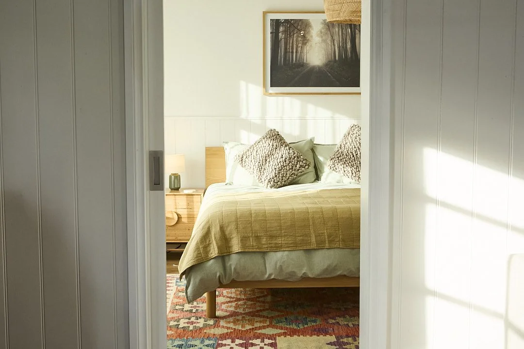 Sunlit bedroom viewed through an open doorway, with a neatly made bed, textured pillows, wood bed-side table, framed forest photograph, and colorful patterned rug.
