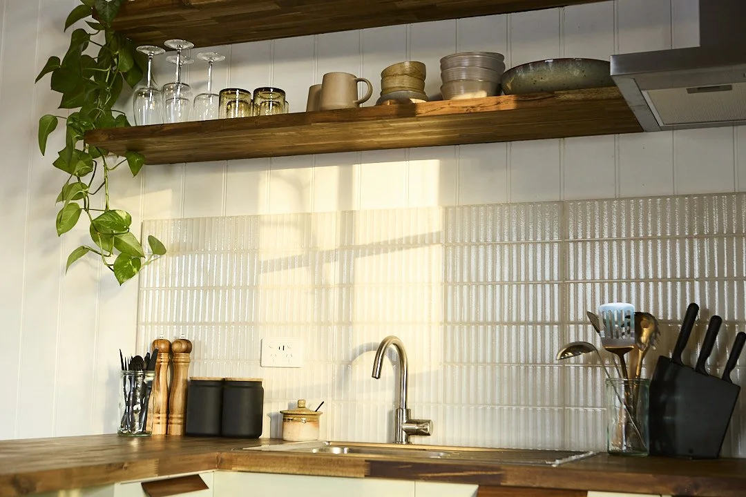 Warm, sunlit kitchen with wooden open shelves, hanging glasses, potted vine plant, tiled backsplash, sink, and cooking utensils