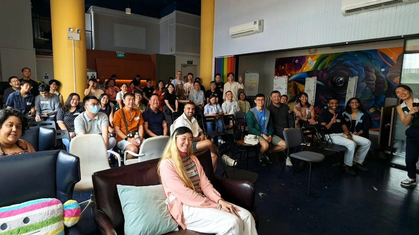 About 40 people seated together at Proud Spaces during the launch of the Queer-Affirming Therapy Code of Practice. Many are smiling towards the camera, while a speaker holding a microphone stands beside a colourful bird mural and rainbow flag.