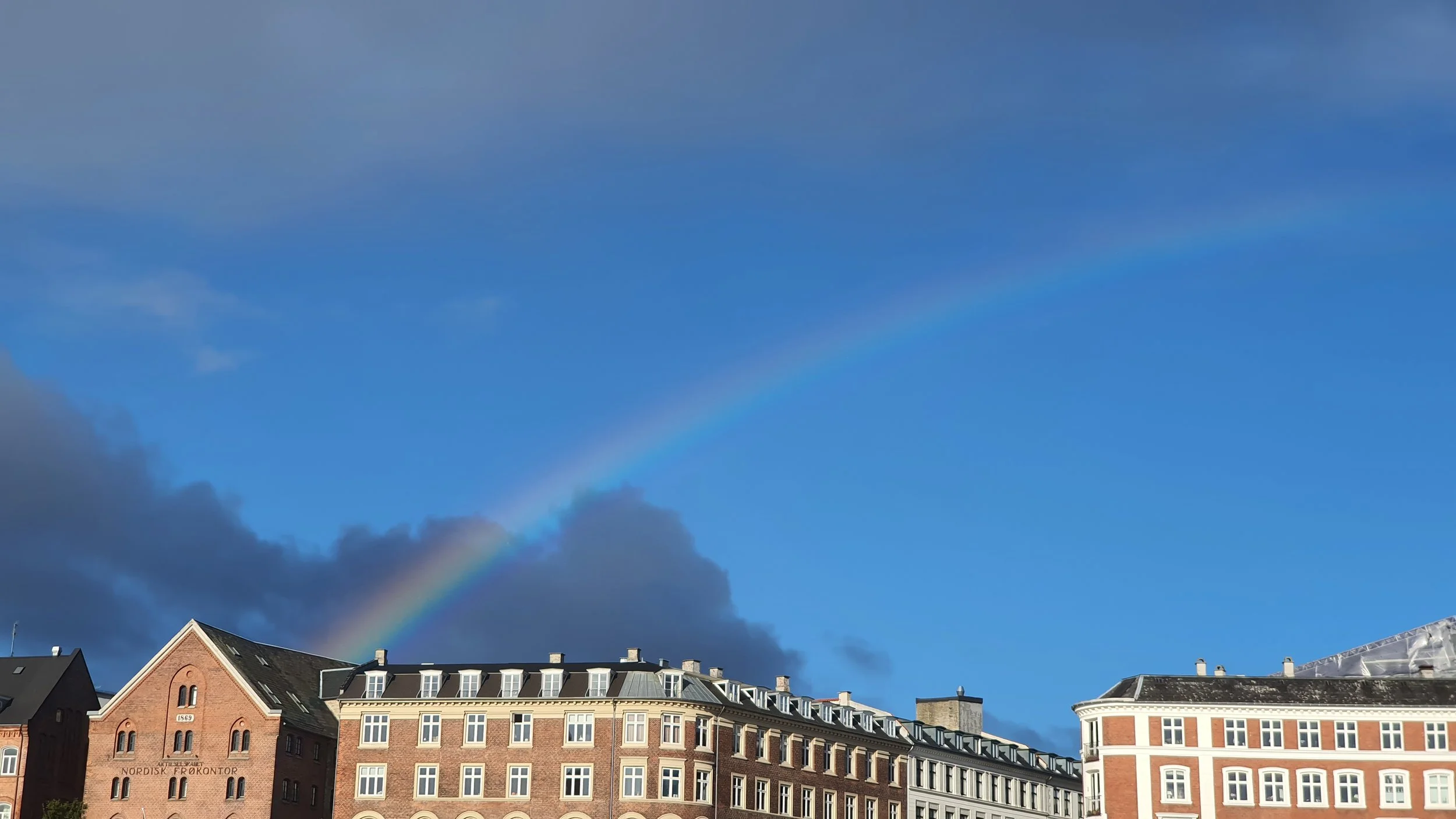 Rainbow arching over a row of brick and stone buildings with a partly cloudy sky.