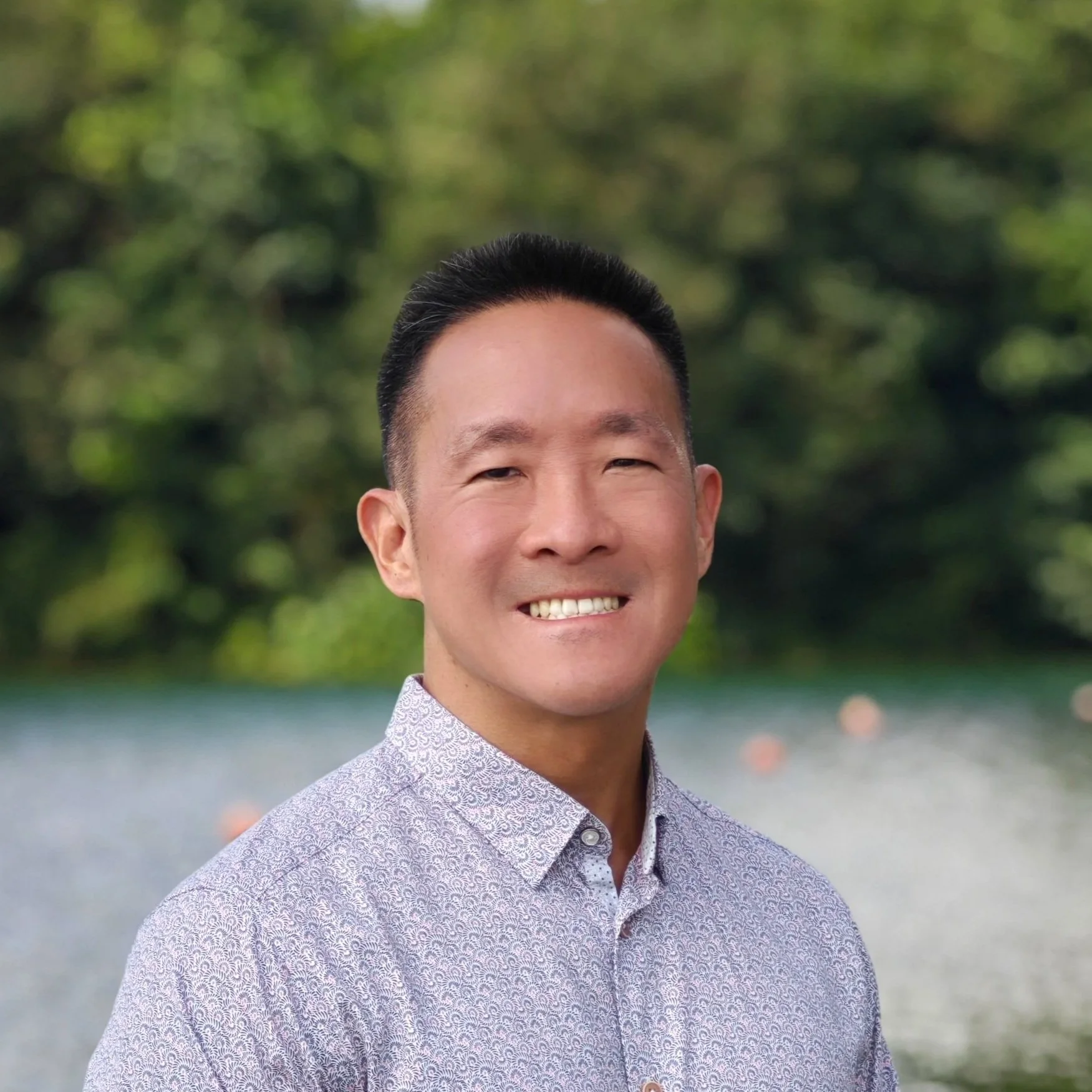 A smiling man stands outdoors in front of a calm lake, wearing a long-sleeve button-down shirt with a subtle patterned design. His hands are clasped in front of him, and the background shows lush green trees under a partly cloudy sky.