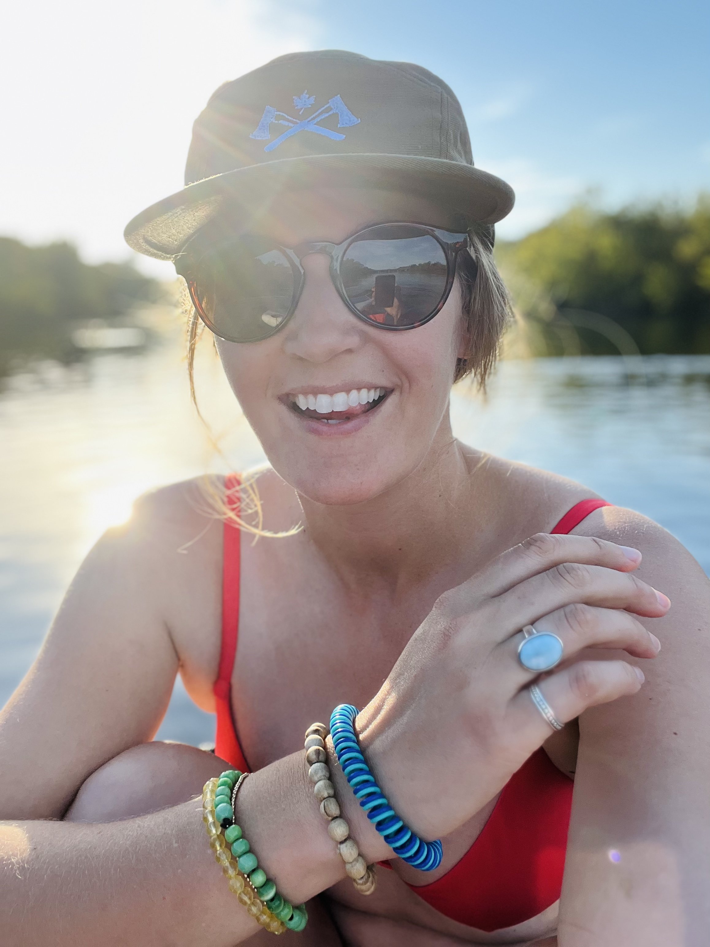 Woman smiling outdoors on a sunny day, wearing large sunglasses, a cap with a blue emblem, a red top, and multiple colorful bracelets, with a body of water and trees in the background.