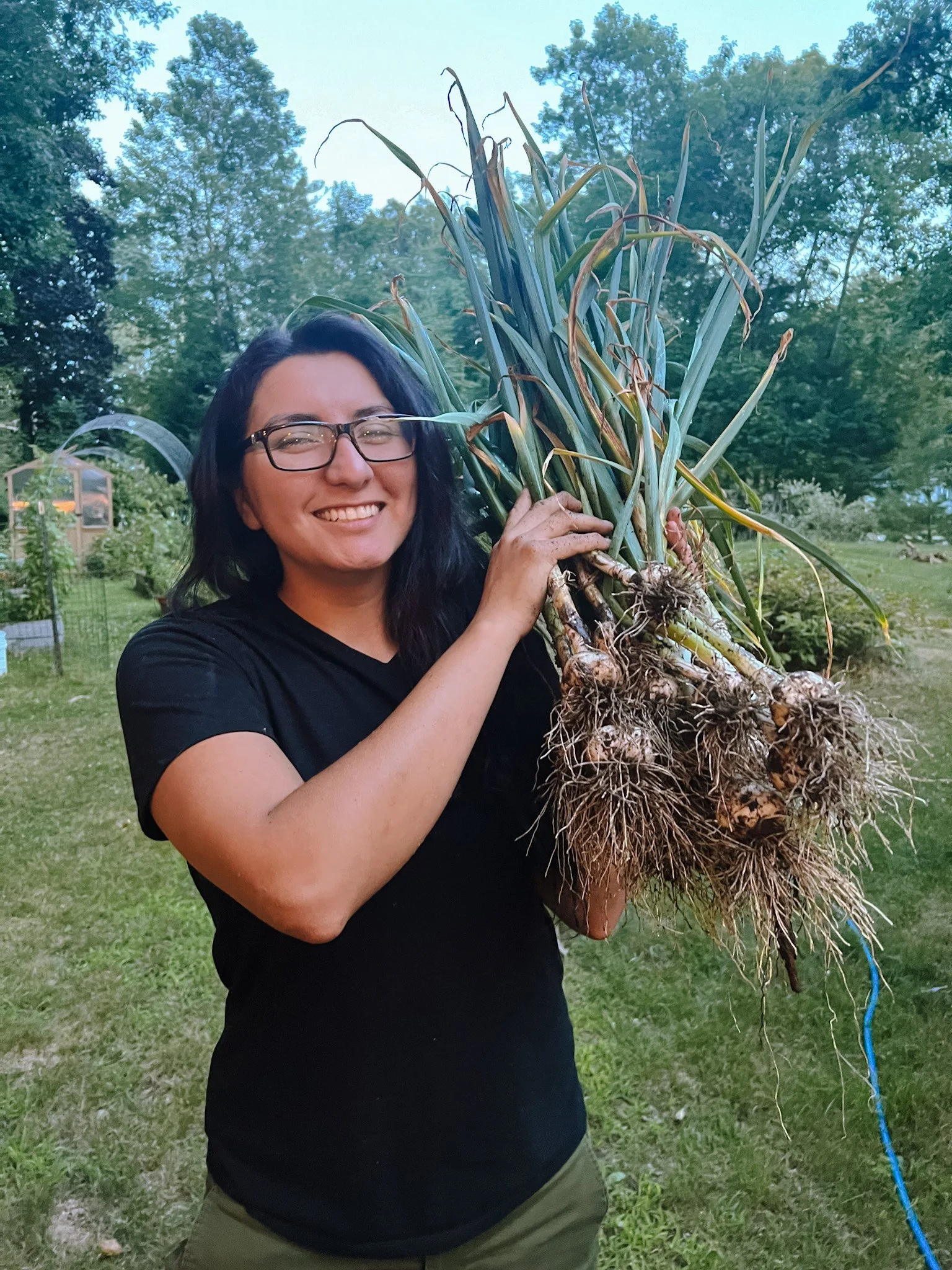 A woman with glasses and a black shirt smiling outdoors while holding a large bunch of freshly harvested garlic bulbs with roots still attached.