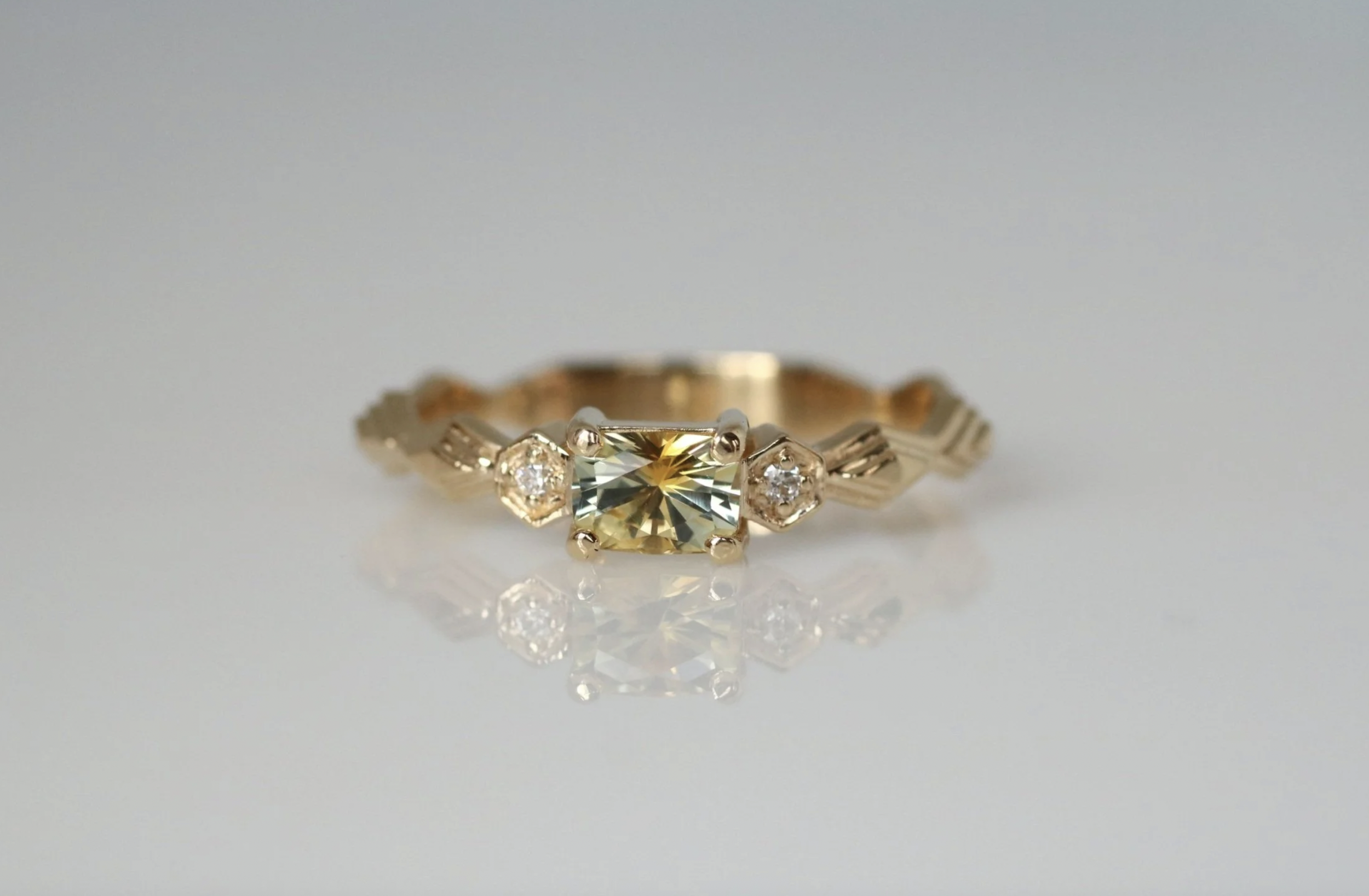 Gold ring with a central yellow gemstone and two smaller clear gemstones on each side, reflecting on a white surface.