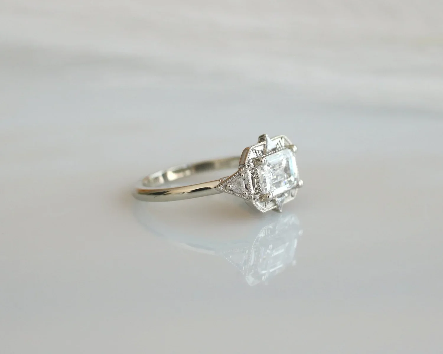 A silver ring with a large central square-cut diamond and smaller diamonds in a geometric setting on a reflective white surface.