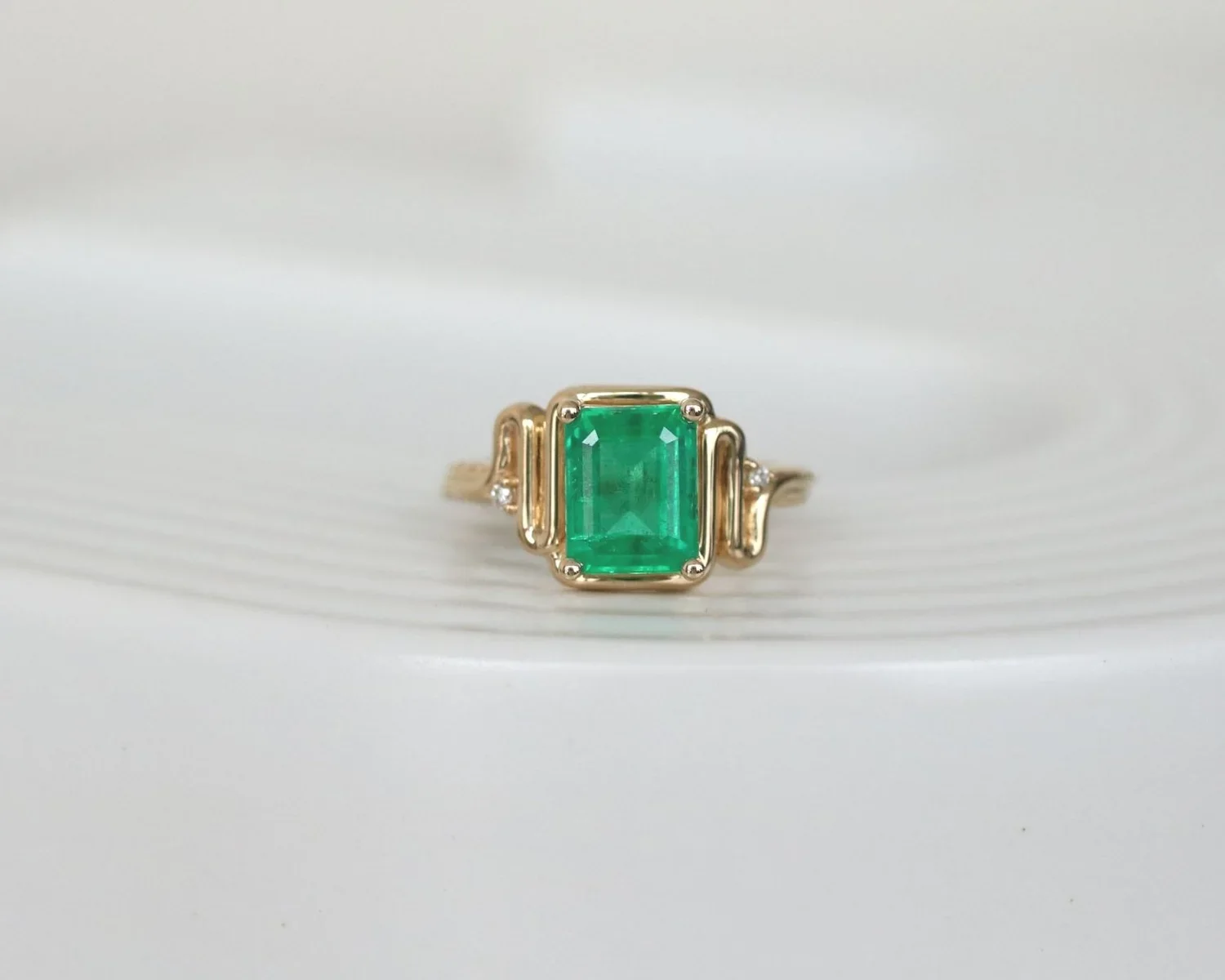 Gold ring with rectangular green emerald gemstone and small diamonds on the sides, on a white surface.