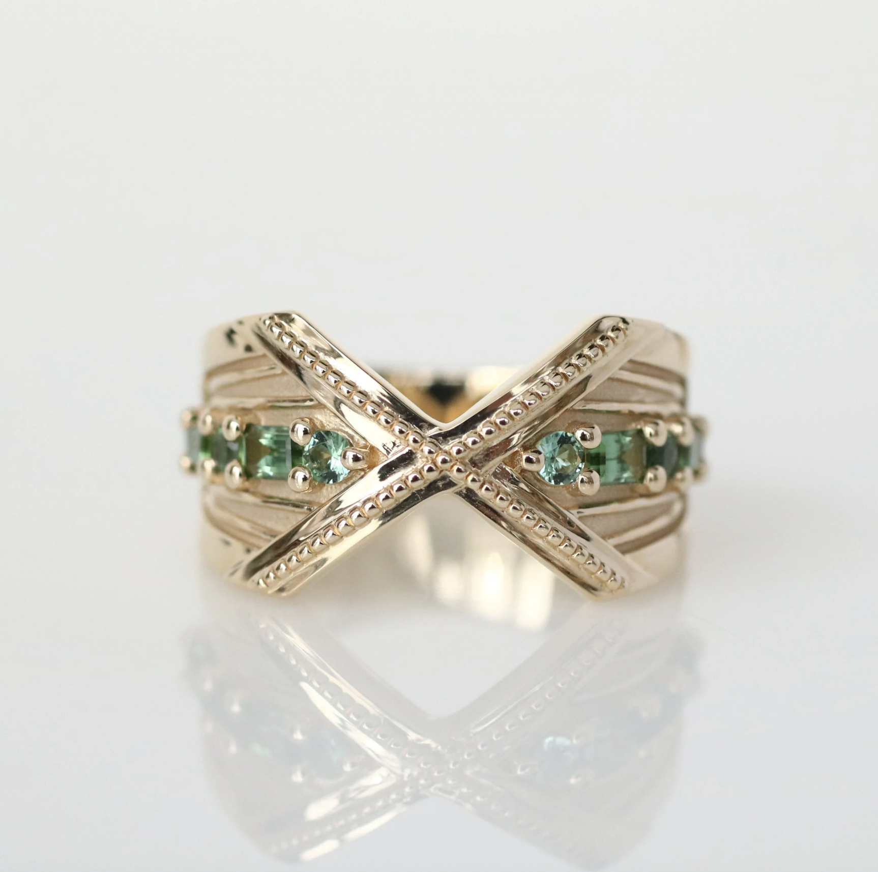Gold ring with crossed band design and embedded green gemstones, reflected on a white surface.