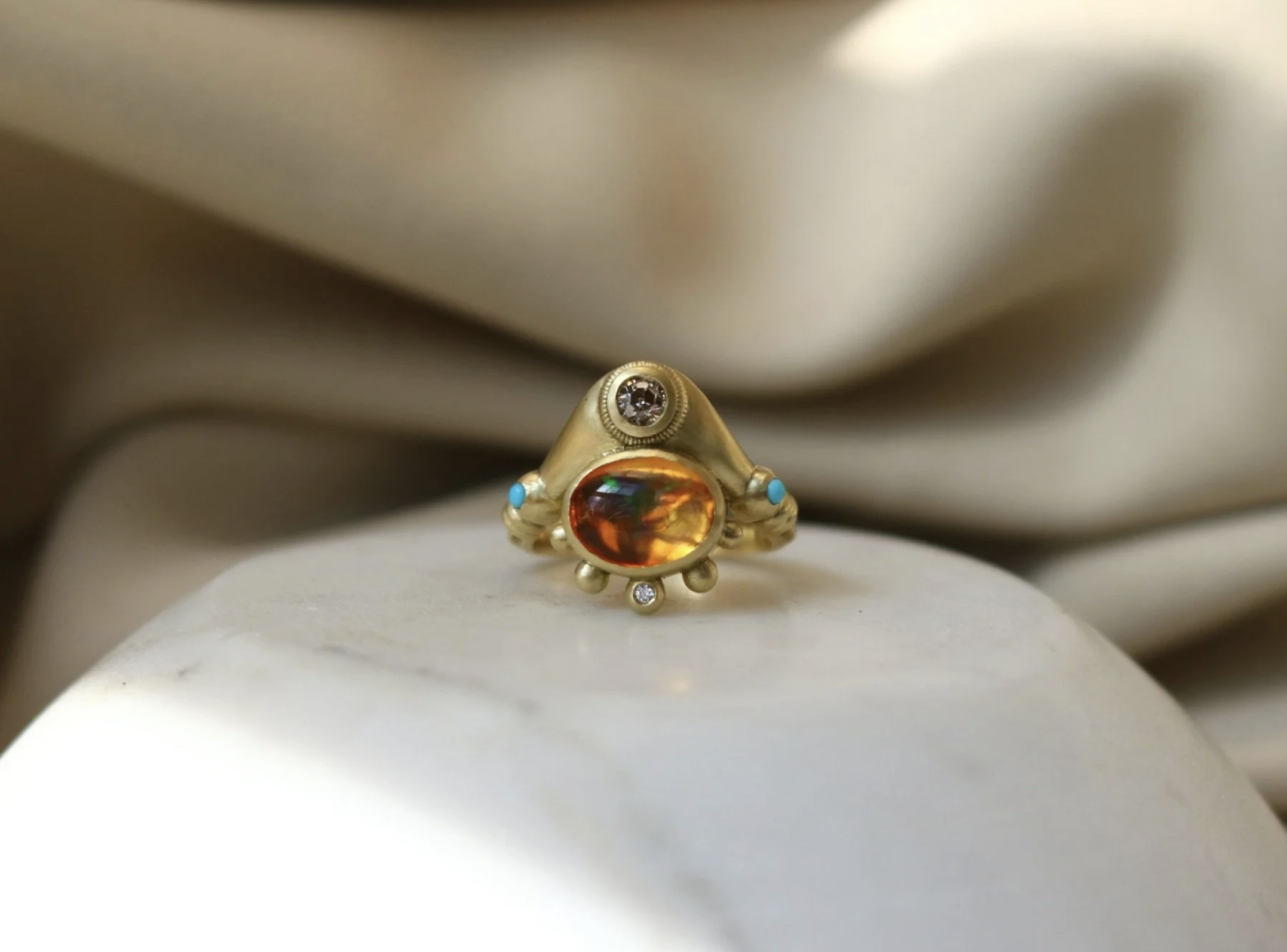 A gold ring with a large amber stone and small turquoise and diamond accents, resting on a smooth white surface with a beige fabric background.