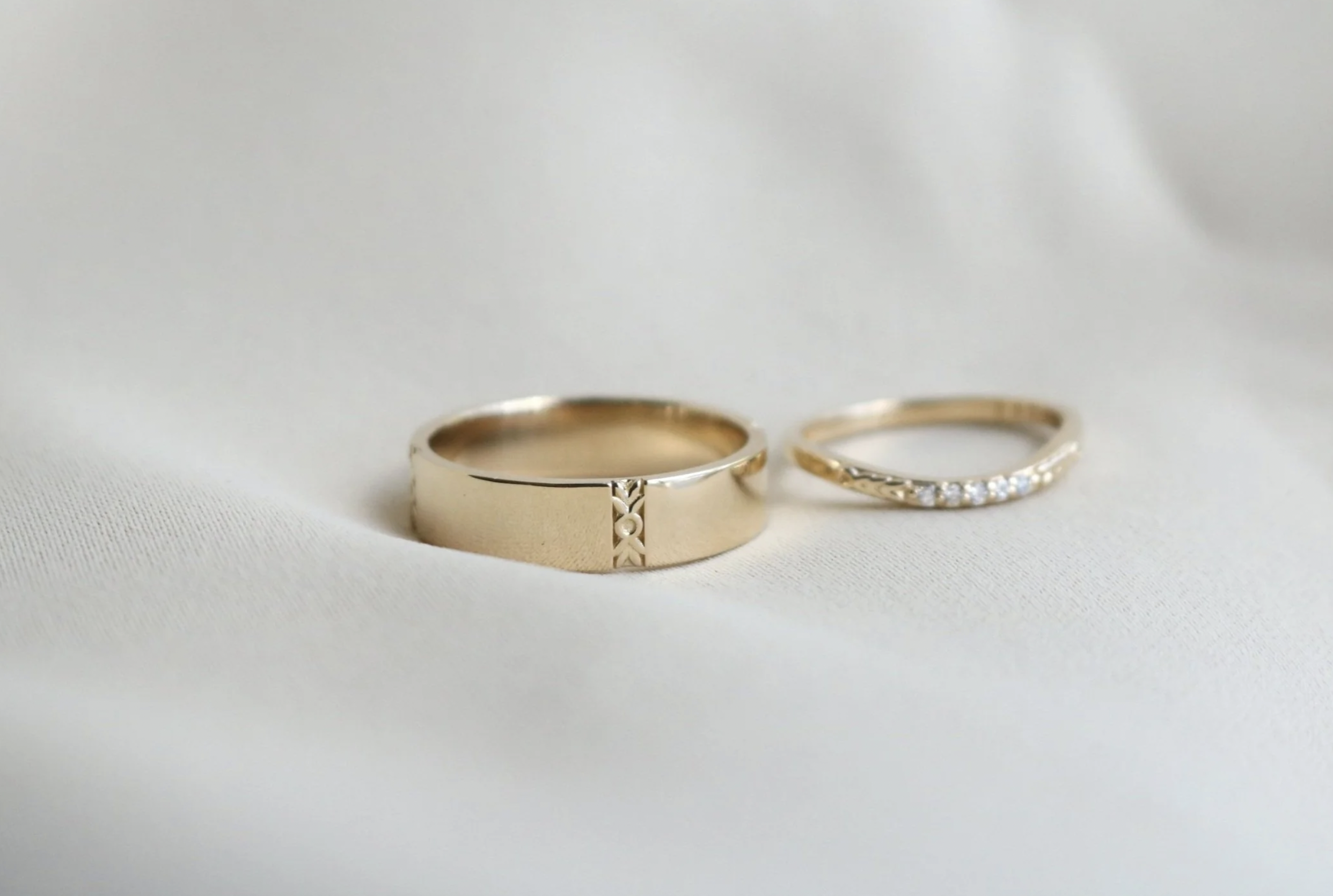 Two gold rings on a white fabric surface, one thick with a engraved design and the other thin with embedded diamonds.