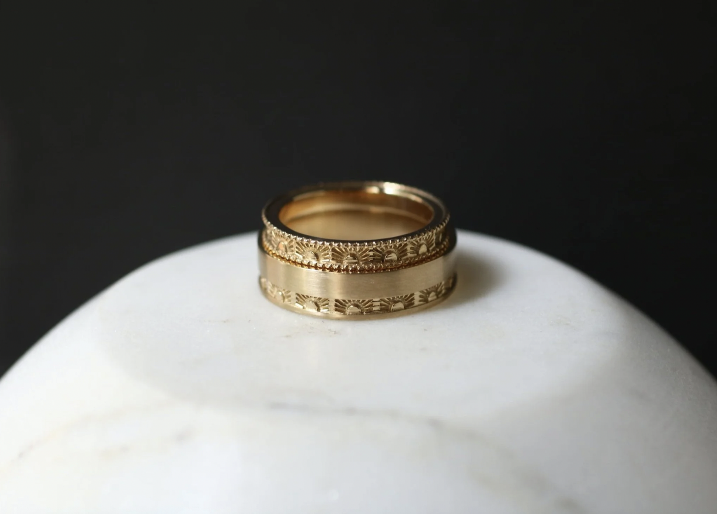 Two gold rings with intricate designs, stacked on top of each other, placed on a white rounded surface against a black background.