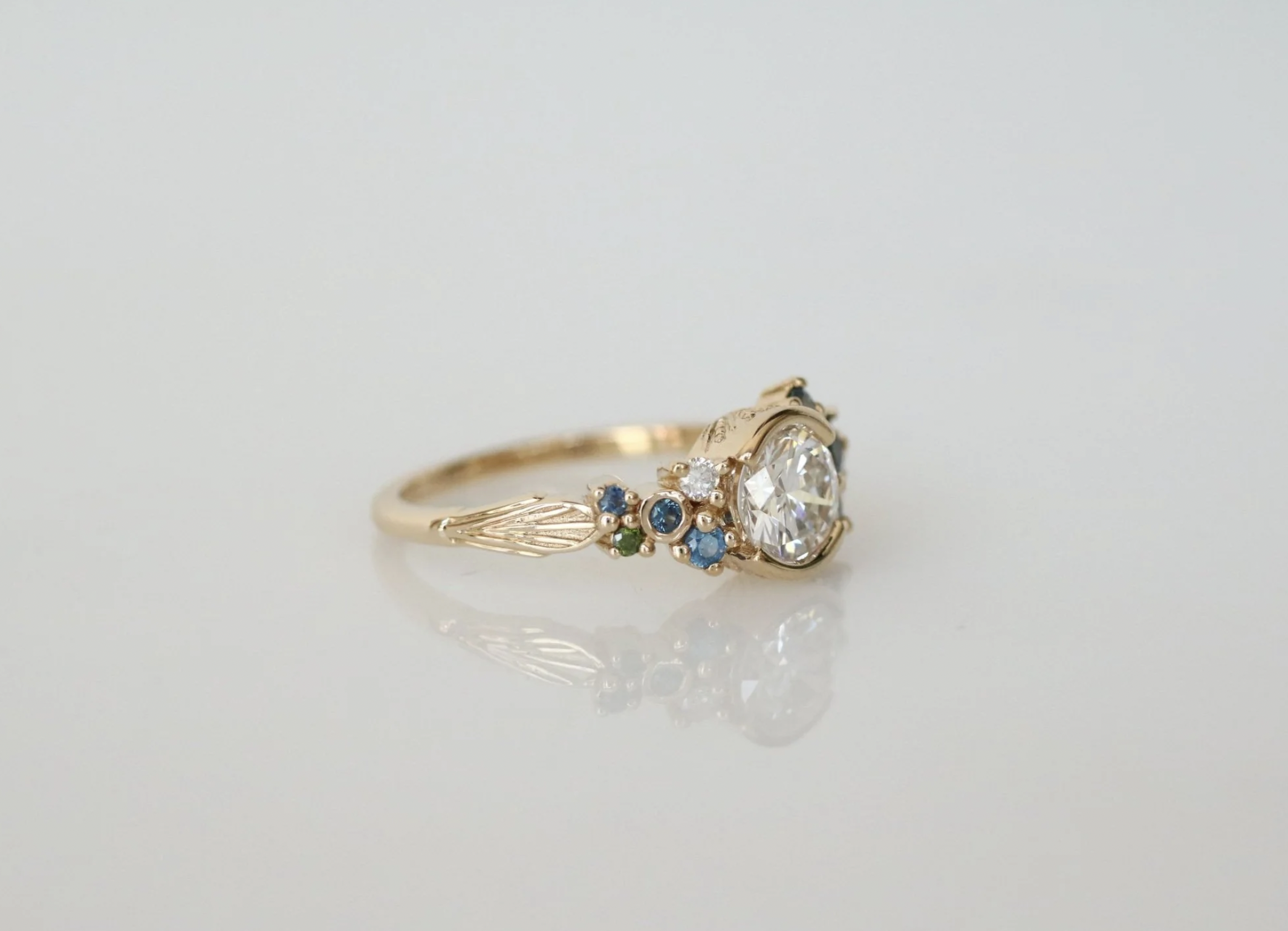 A gold ring with a central clear gemstone and smaller blue, green, and clear gemstones on the band, placed on a white surface.