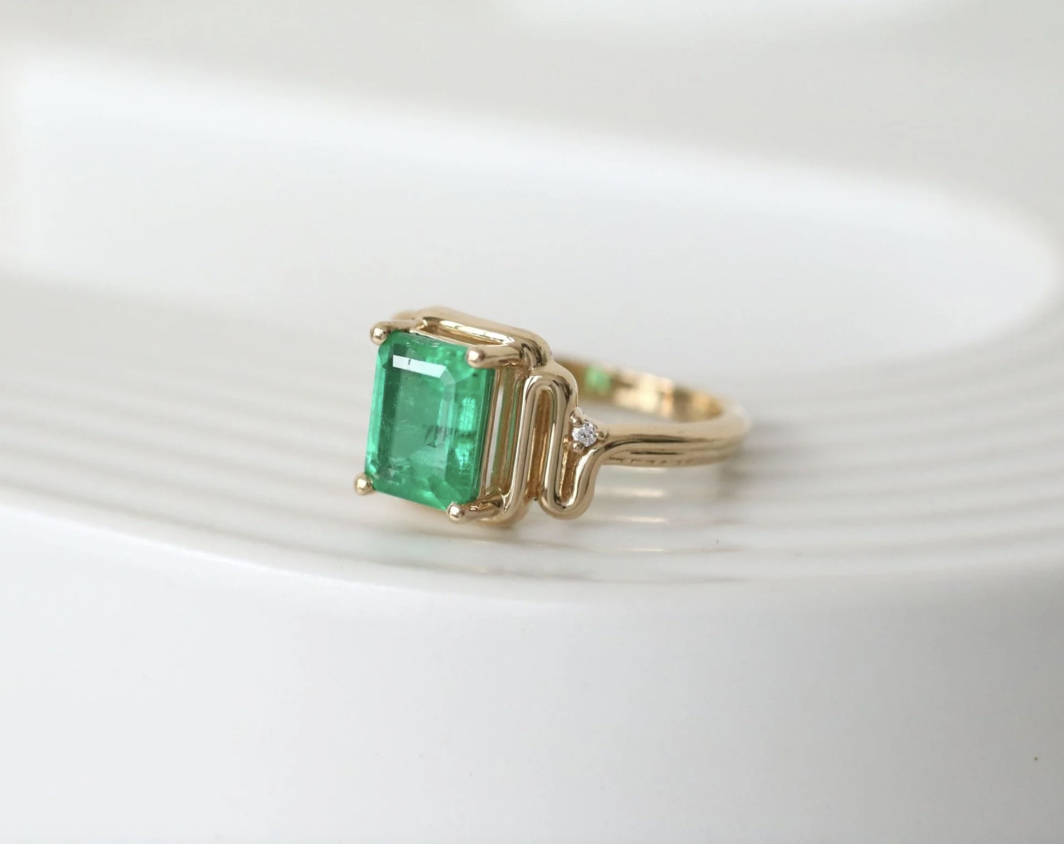 Gold ring with a large square emerald gemstone and small diamonds, resting on a white textured surface.