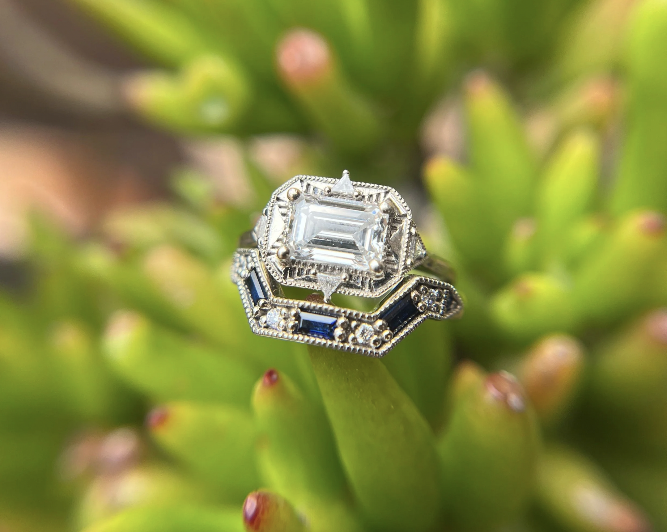 A silver ring with an emerald-cut clear gemstone and blue accents, resting on a green plant with small red-tipped buds.