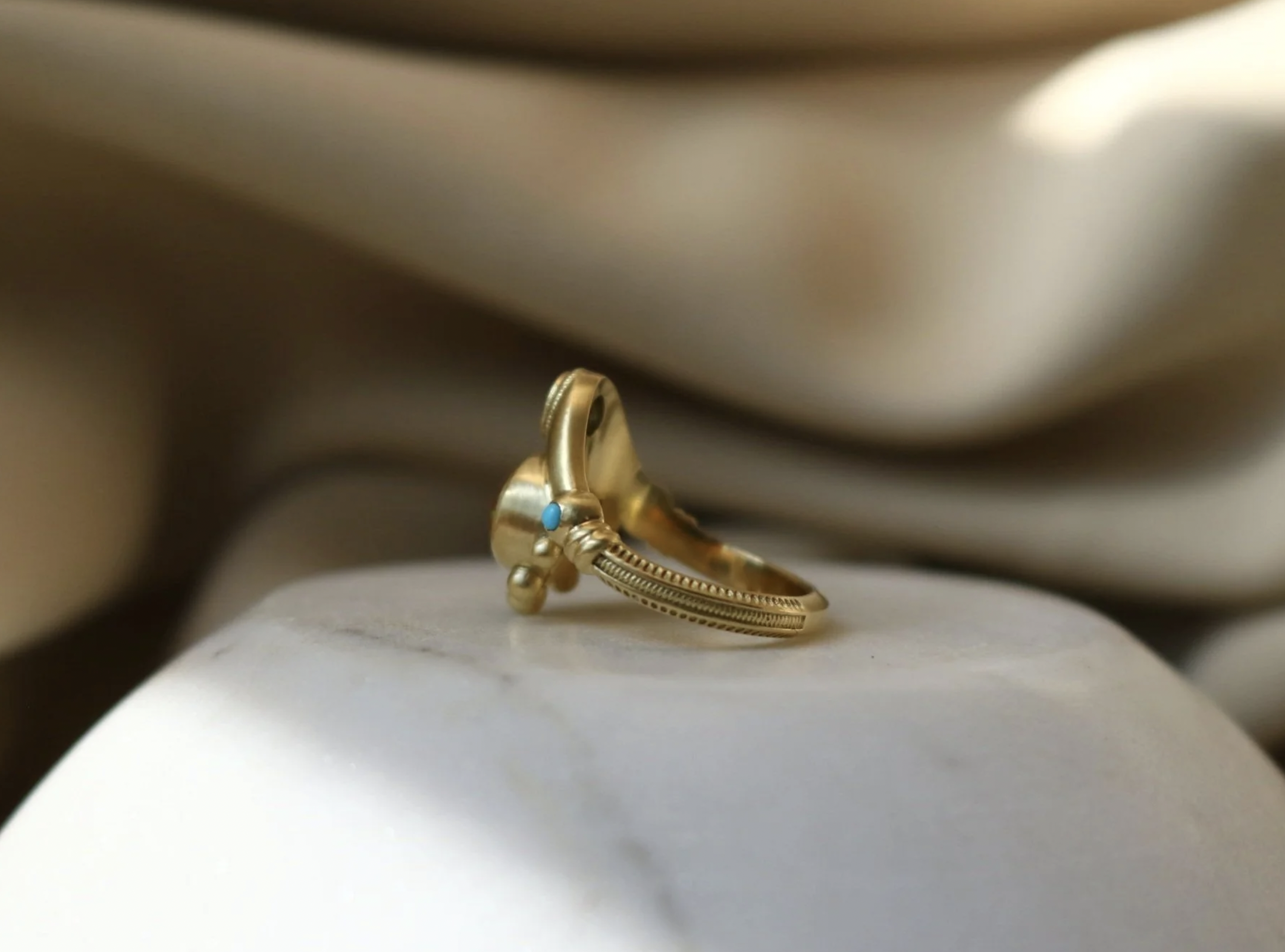 Close-up of a gold ring with a tiny turquoise stone, resting on a smooth white surface with blurred background.