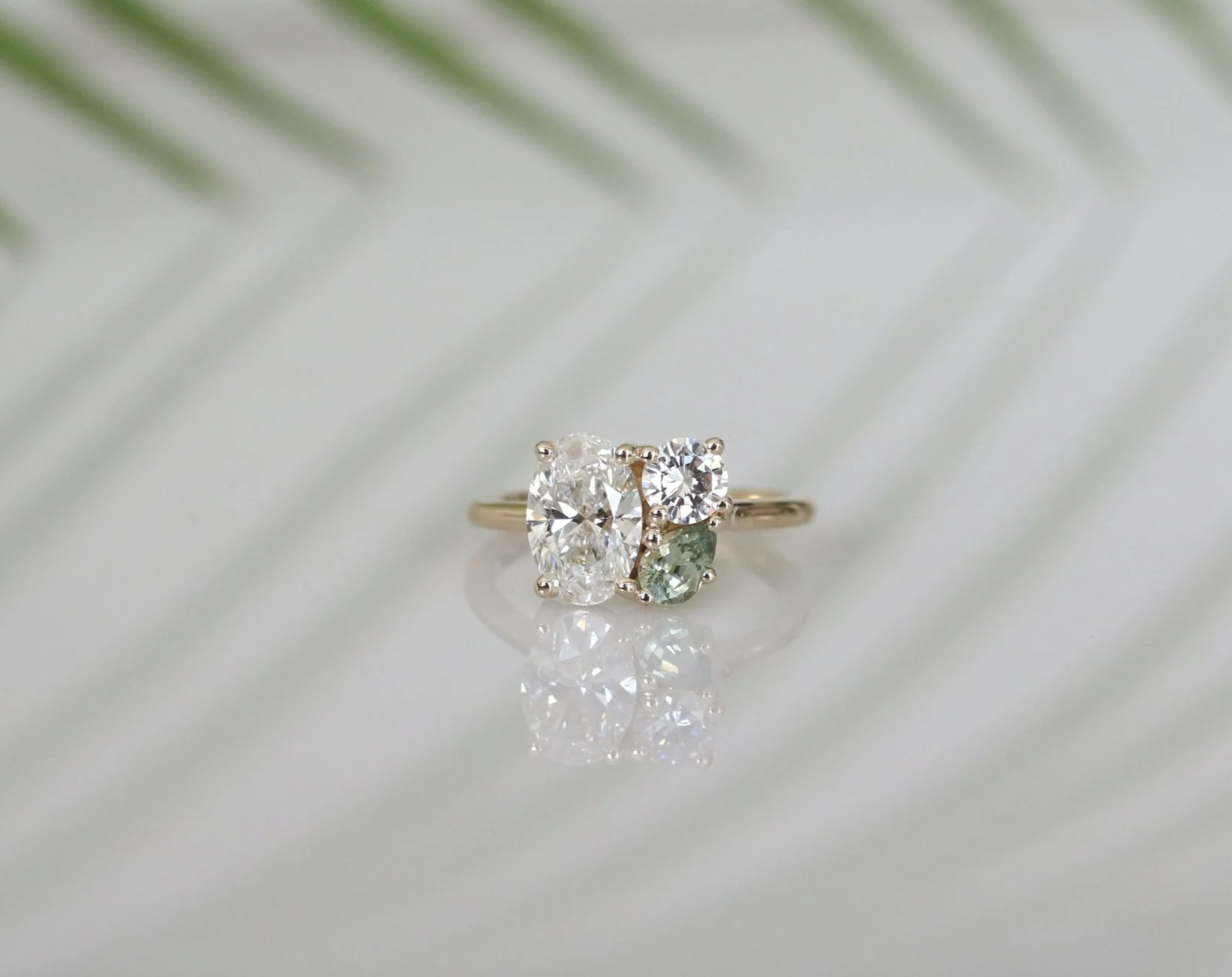 A gold ring with three different sized diamonds and a green gemstone, placed on a white surface with a blurred green leaf in the background.