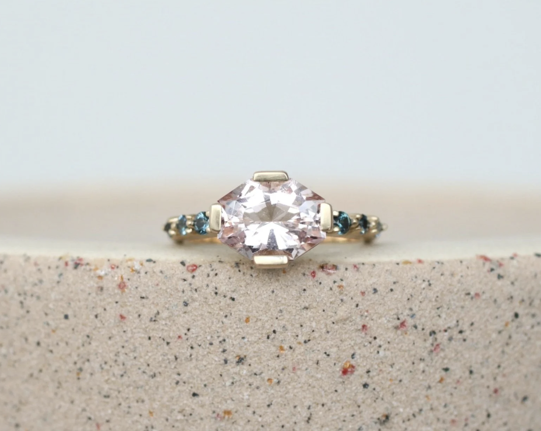 Close-up of a gold ring with a large pinkish clear gemstone and small blue stones on each side, placed on a multi-colored speckled surface.
