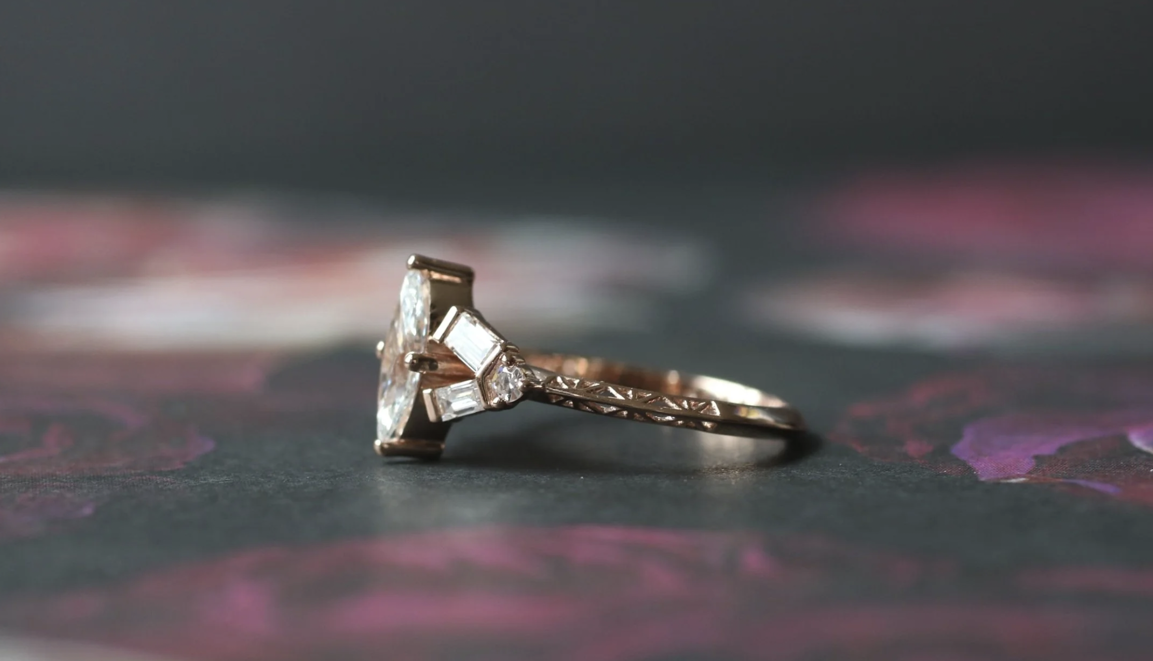 A ring with a large central gemstone, surrounded by smaller gemstones, resting on a surface with pink and purple floral patterns.