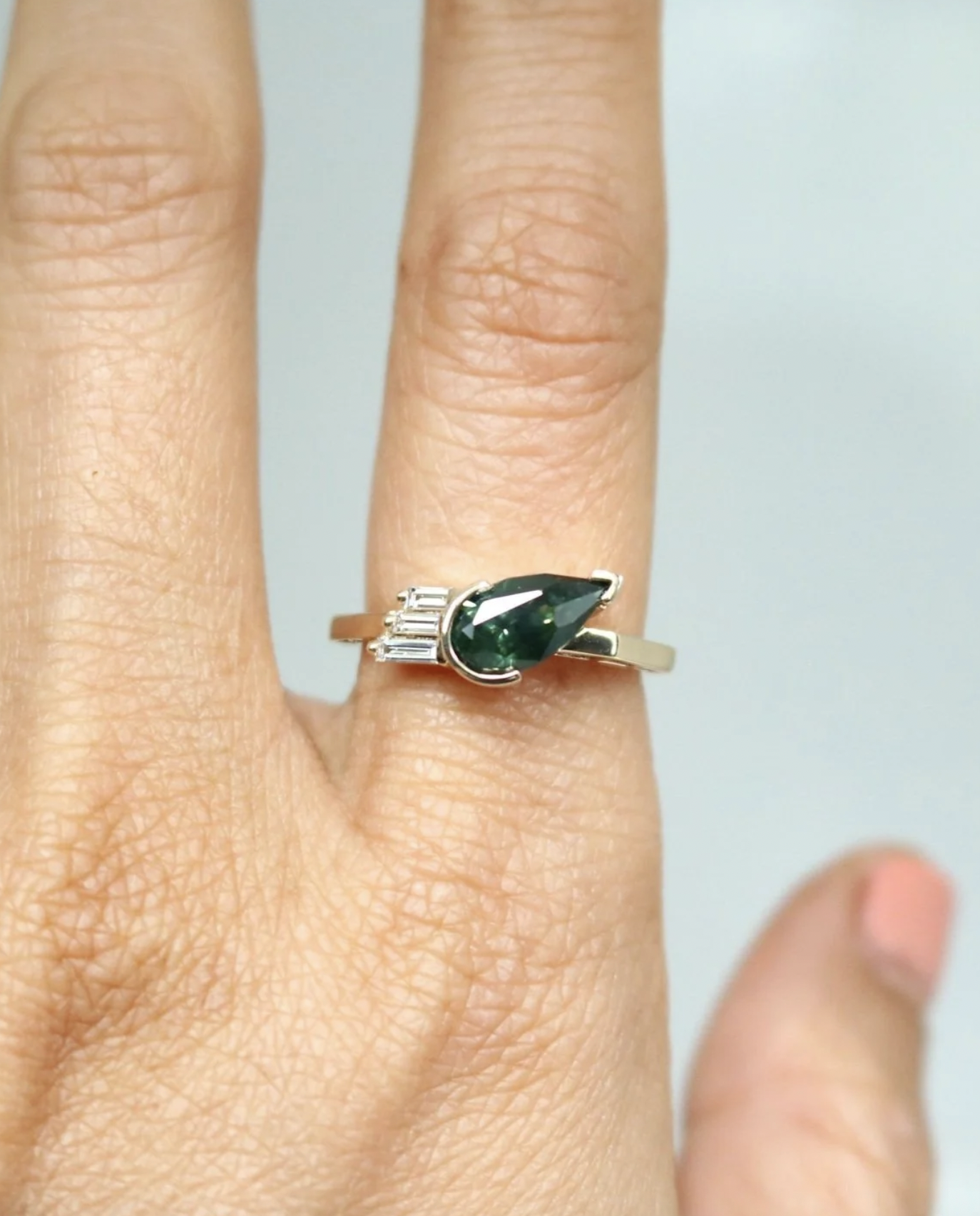 A silver ring with a dark green teardrop-shaped gemstone and three small clear rectangular stones on the side, worn on a person's finger.