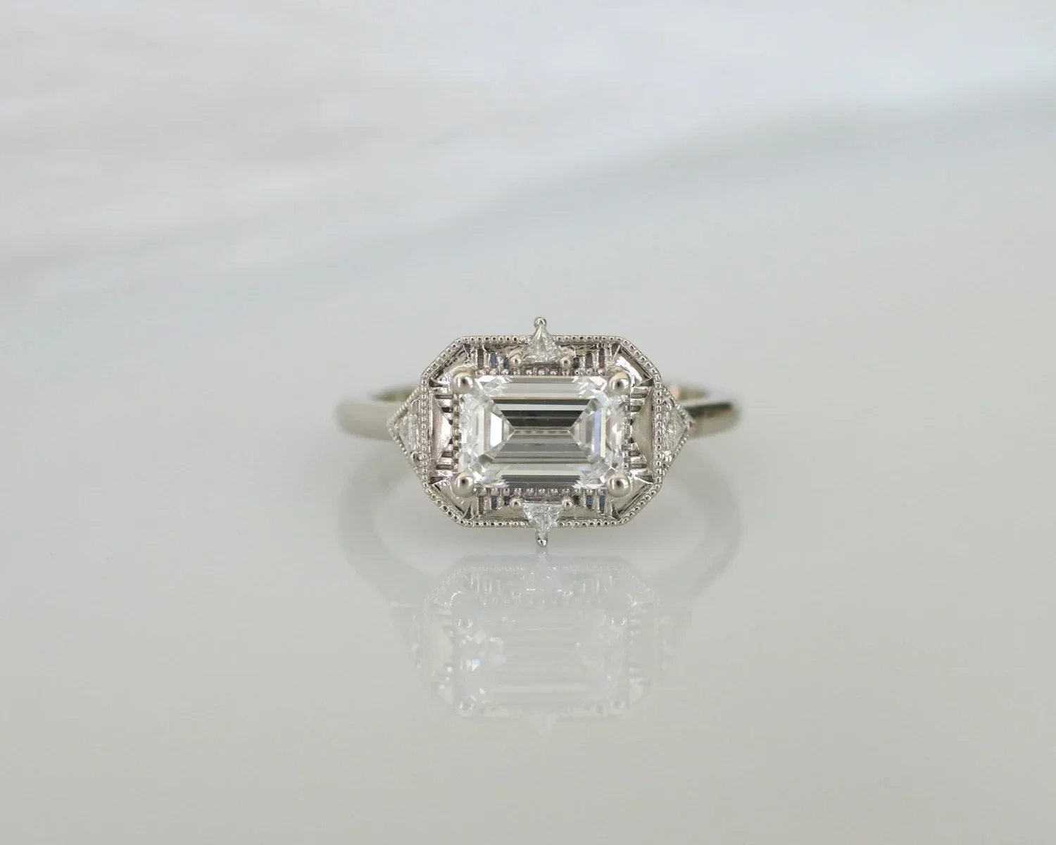Silver ring with an emerald-cut diamond center stone and intricate milgrain and geometric design details.