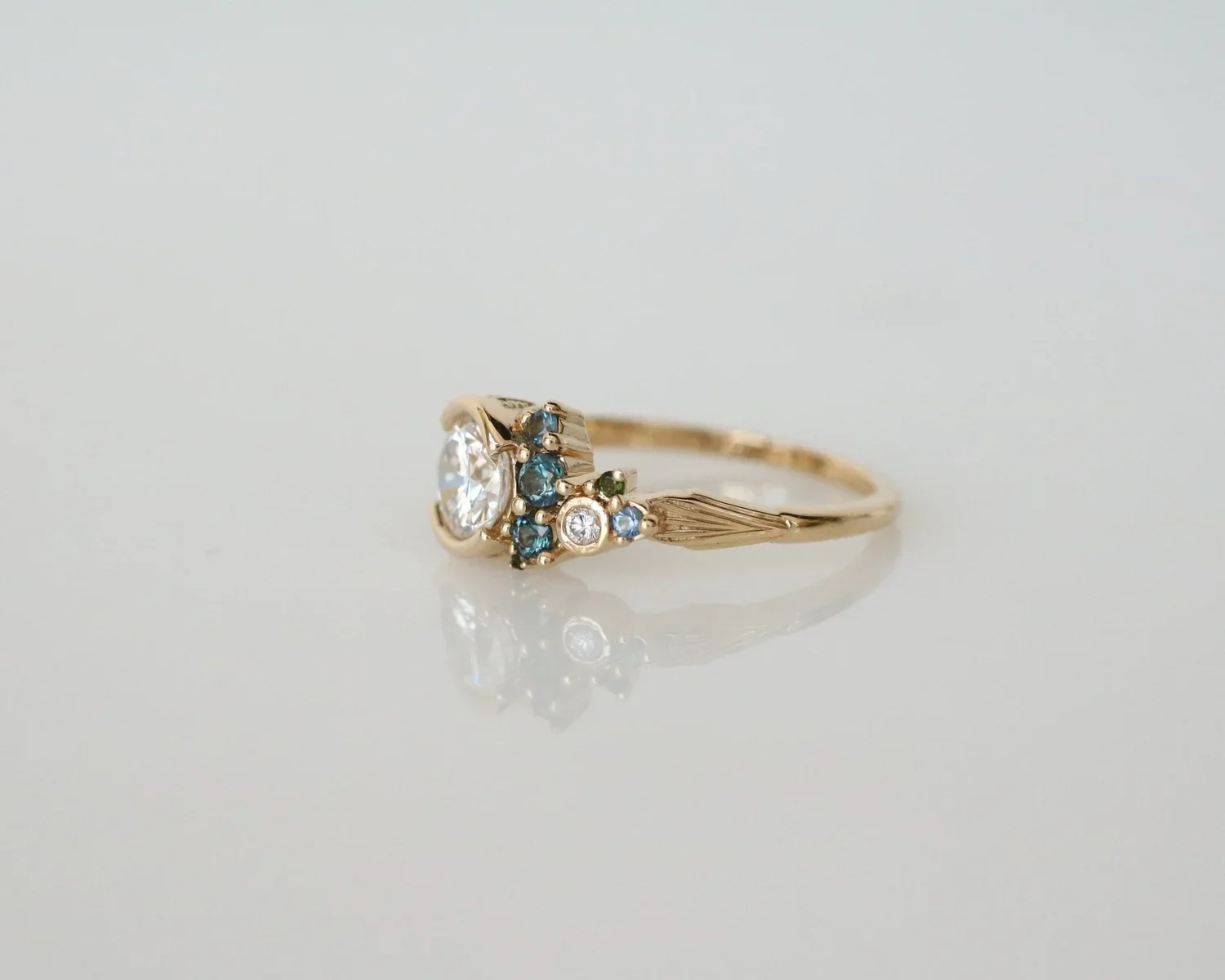 A gold ring with a large central clear gemstone, surrounded by small blue, green, and white gemstones, set on a reflective white surface.