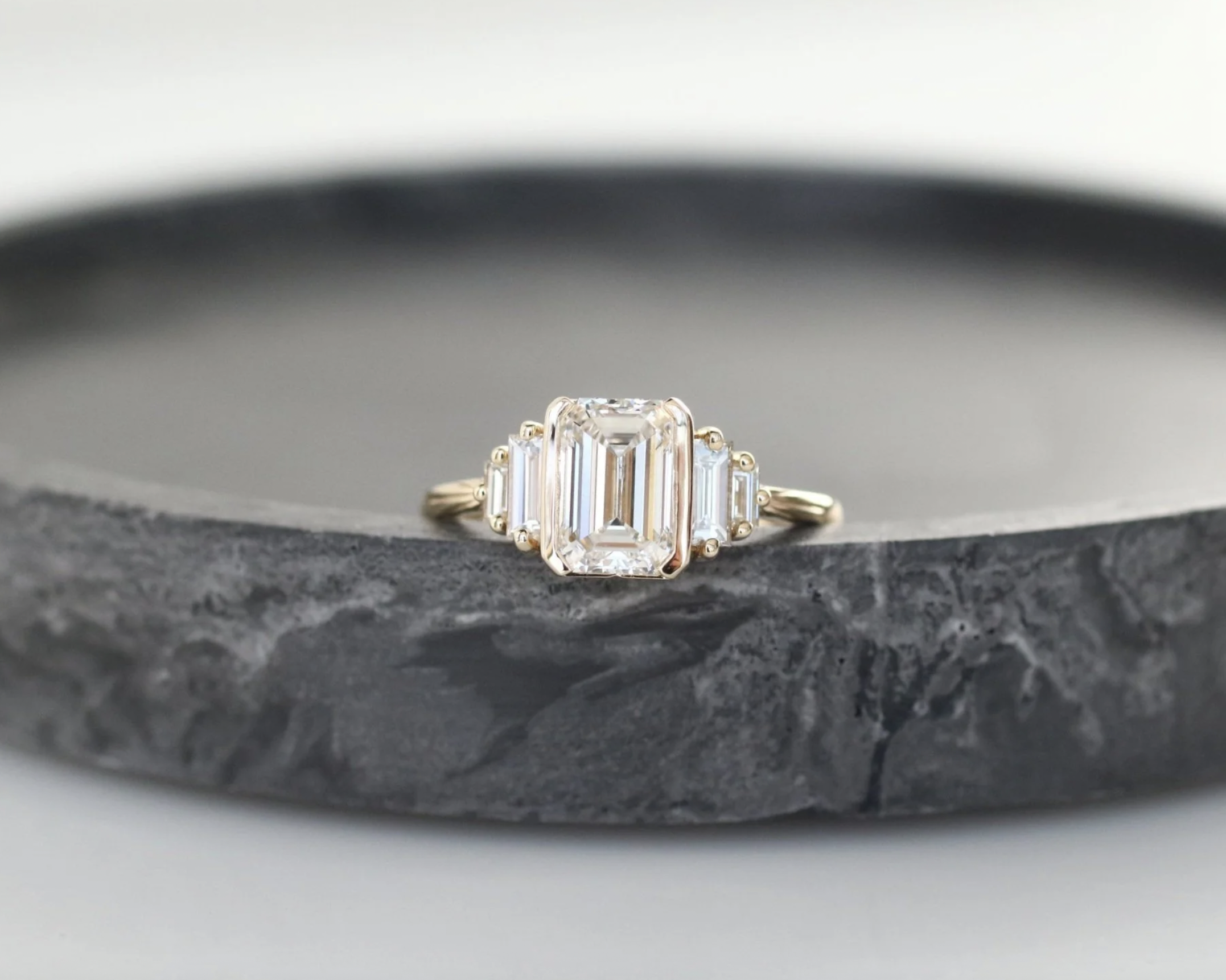 Close-up of a gold ring with an emerald-cut diamond and side baguette diamonds, placed on a dark gray textured surface.