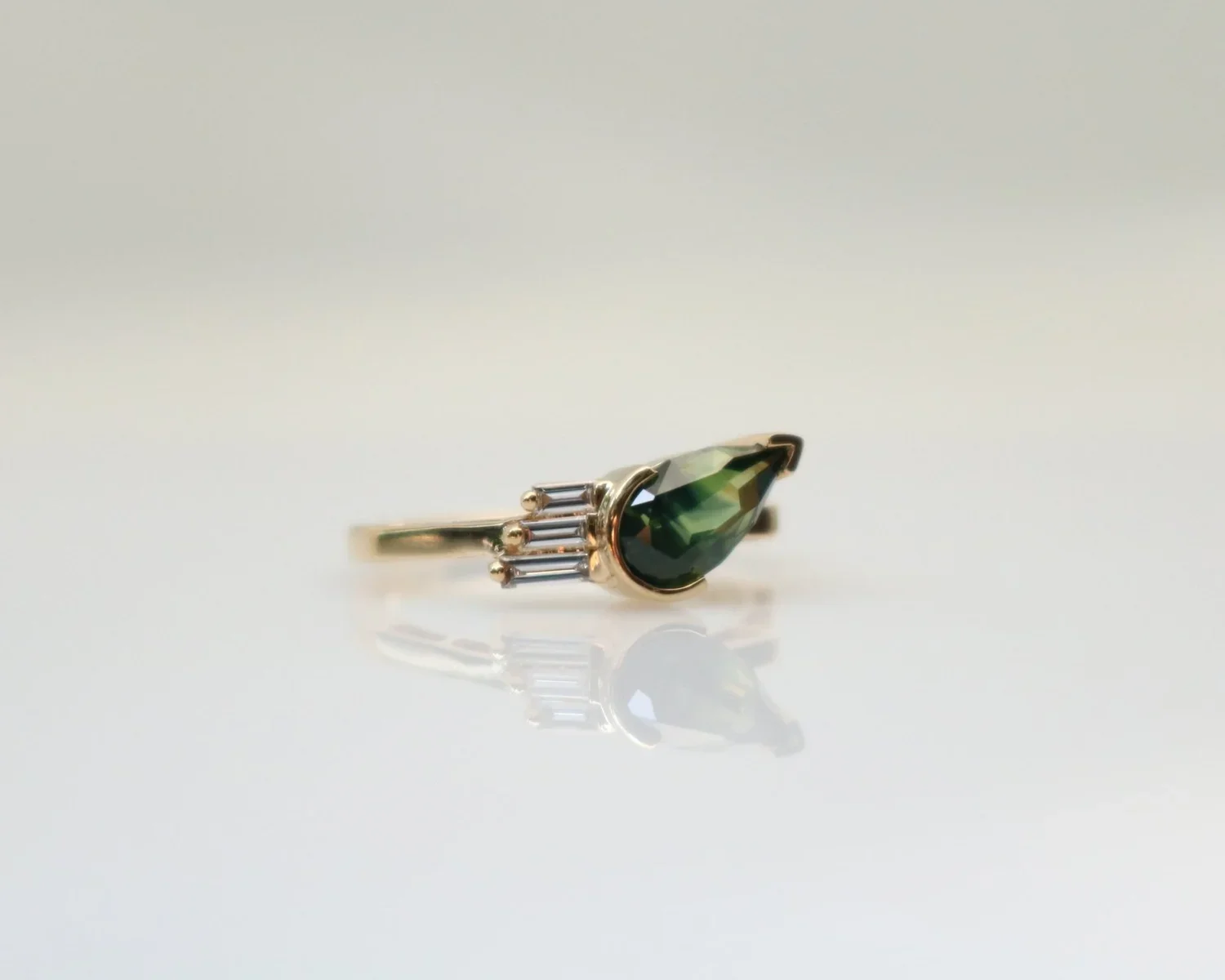 A gold ring with a large green gemstone and three smaller clear gemstones on the side, resting on a reflective surface.