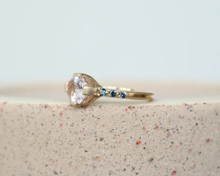 Gold ring with a large central gemstone and three smaller blue gemstones on the band, resting on a speckled surface.