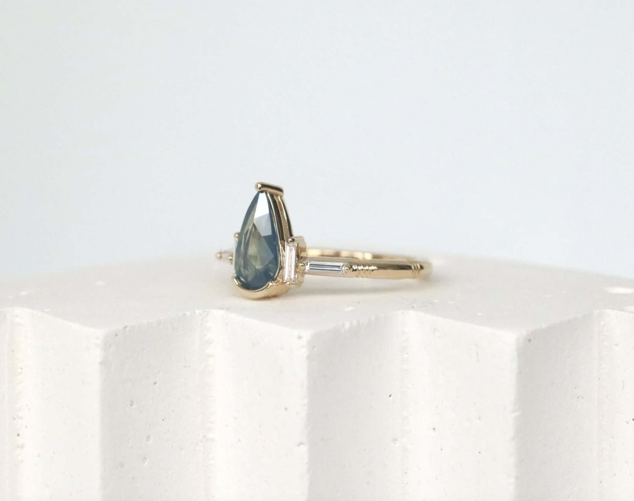 A gold ring with a teardrop-shaped blue gemstone and small baguette-cut clear gemstones on either side, resting on a white textured surface.