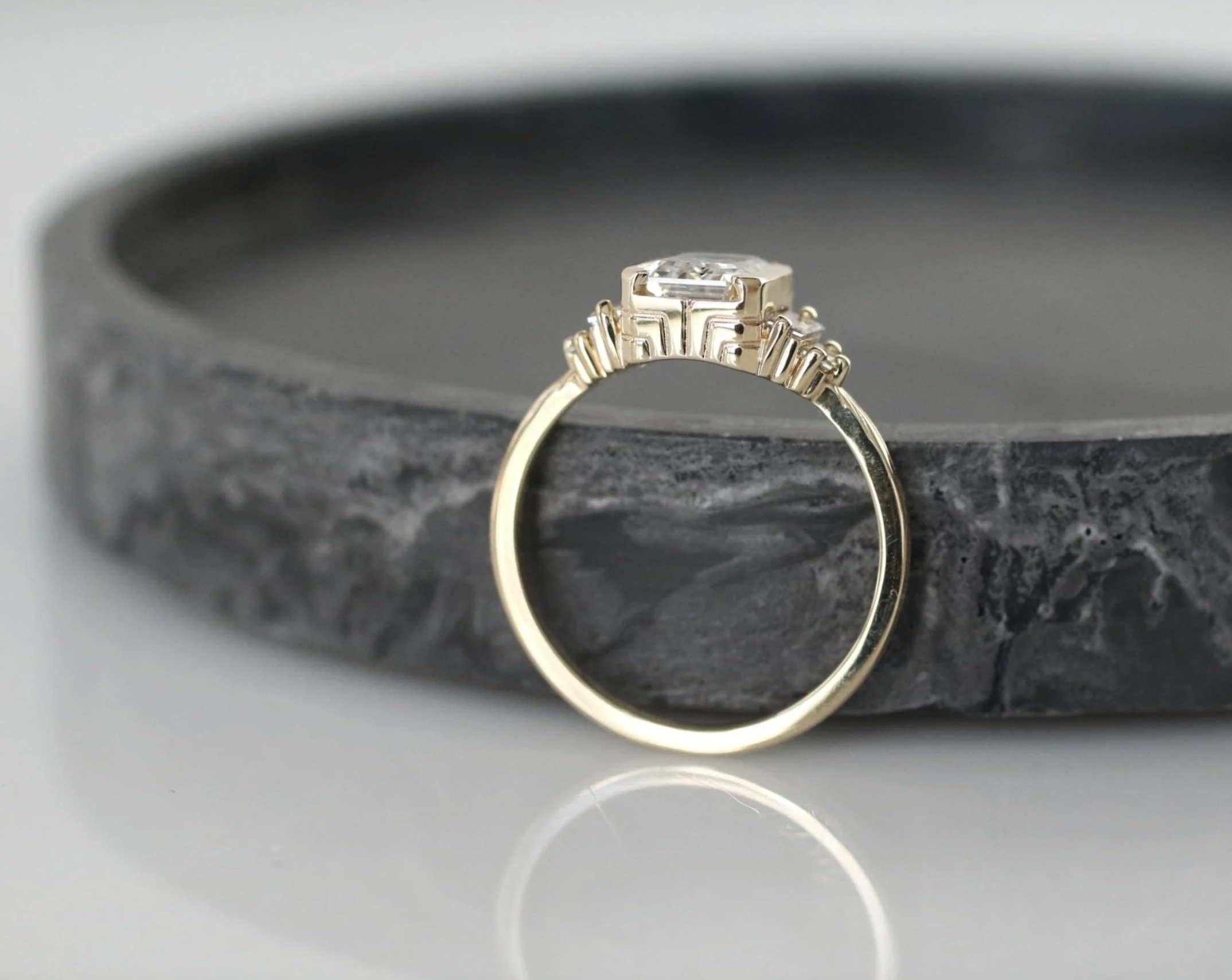A gold ring with a geometric design and a large central gemstone resting on a dark, textured stone surface.