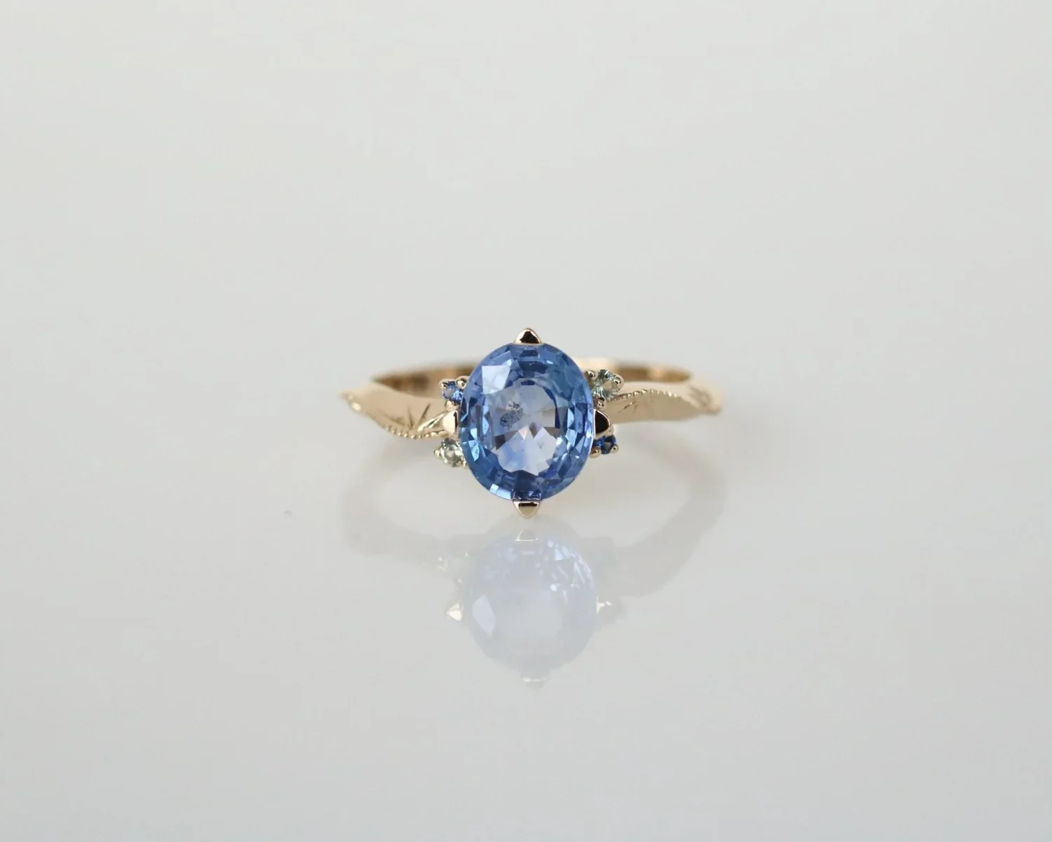 Gold ring with a large oval blue gemstone and small accent stones on each side, reflected on a smooth reflective surface.