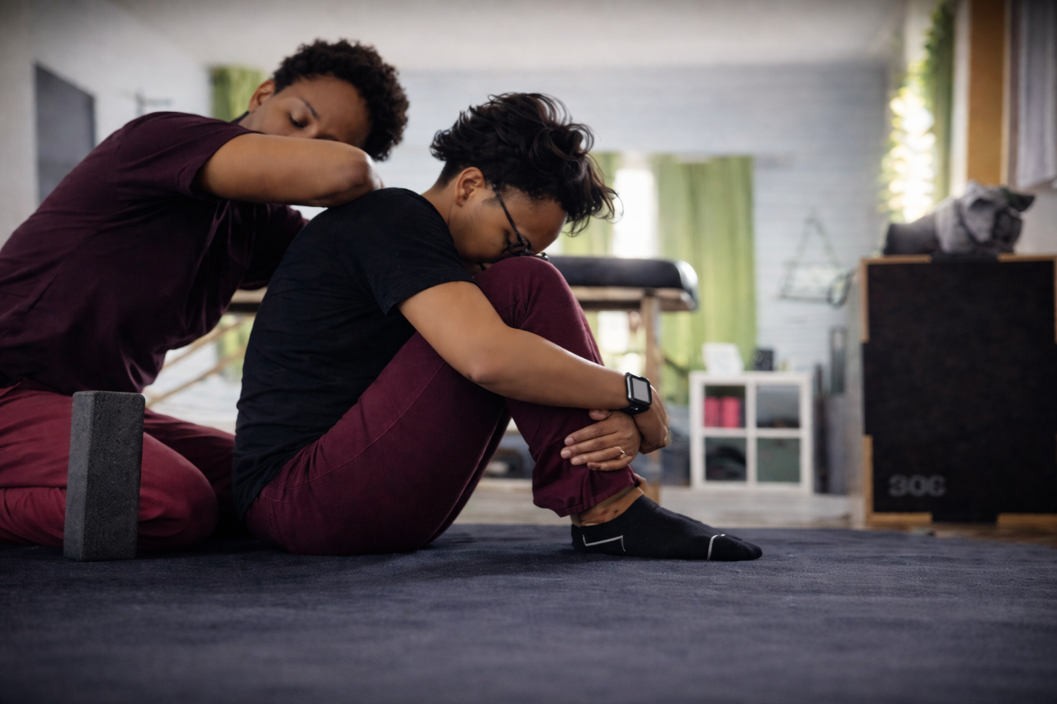 A woman assisting another woman in stretching during a workout in a gym, with a yoga block nearby.