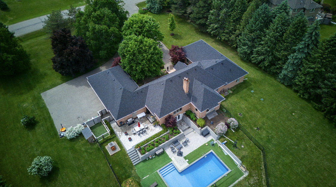 Aerial view of a large backyard with a swimming pool, patio area with outdoor furniture, and lush green trees surrounding a brick house.