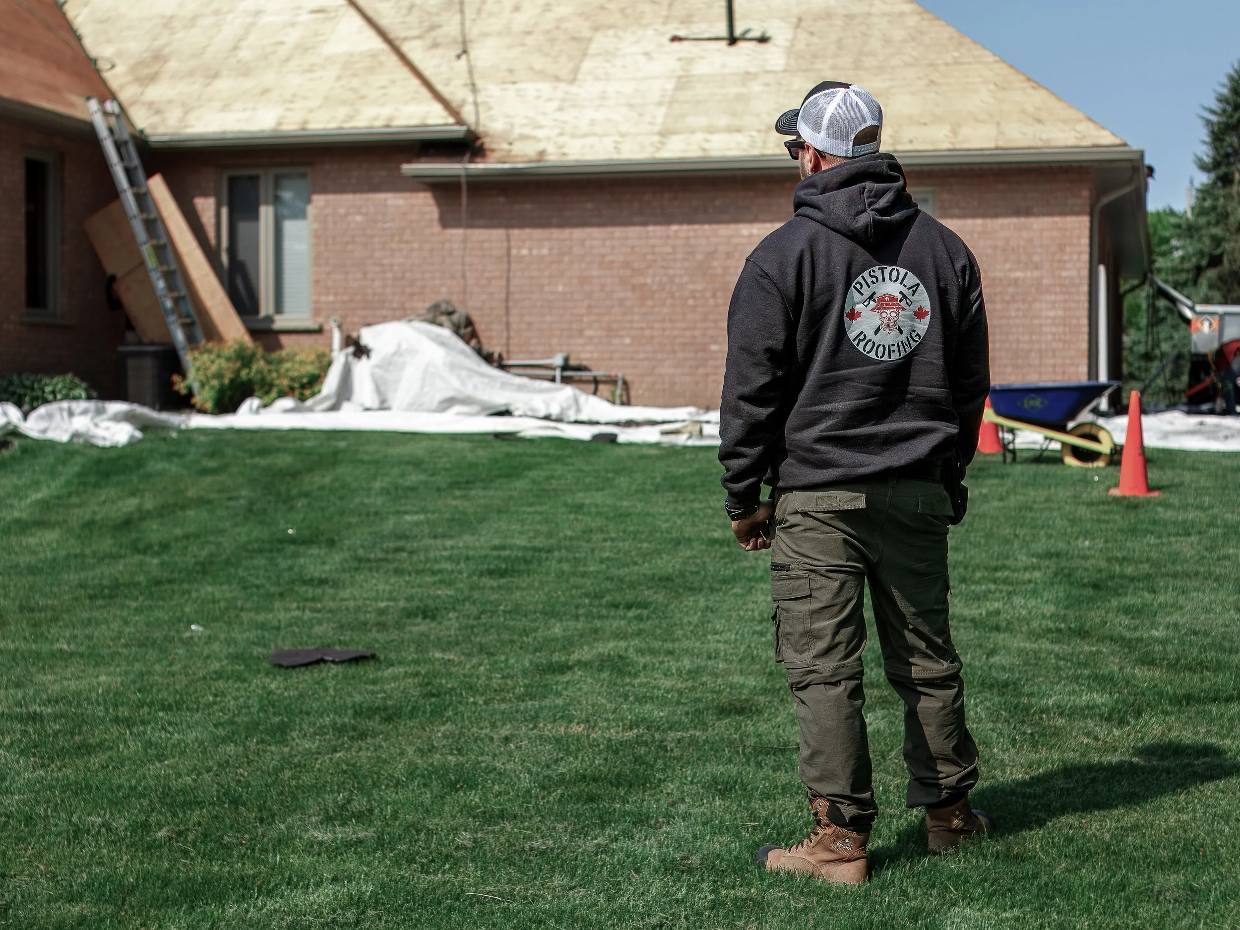 Man wearing a black hoodie with the logo "Pistola Roofing" on the back, standing on a lawn and looking at a house with damage to the roof, surrounded by construction materials and orange cones.