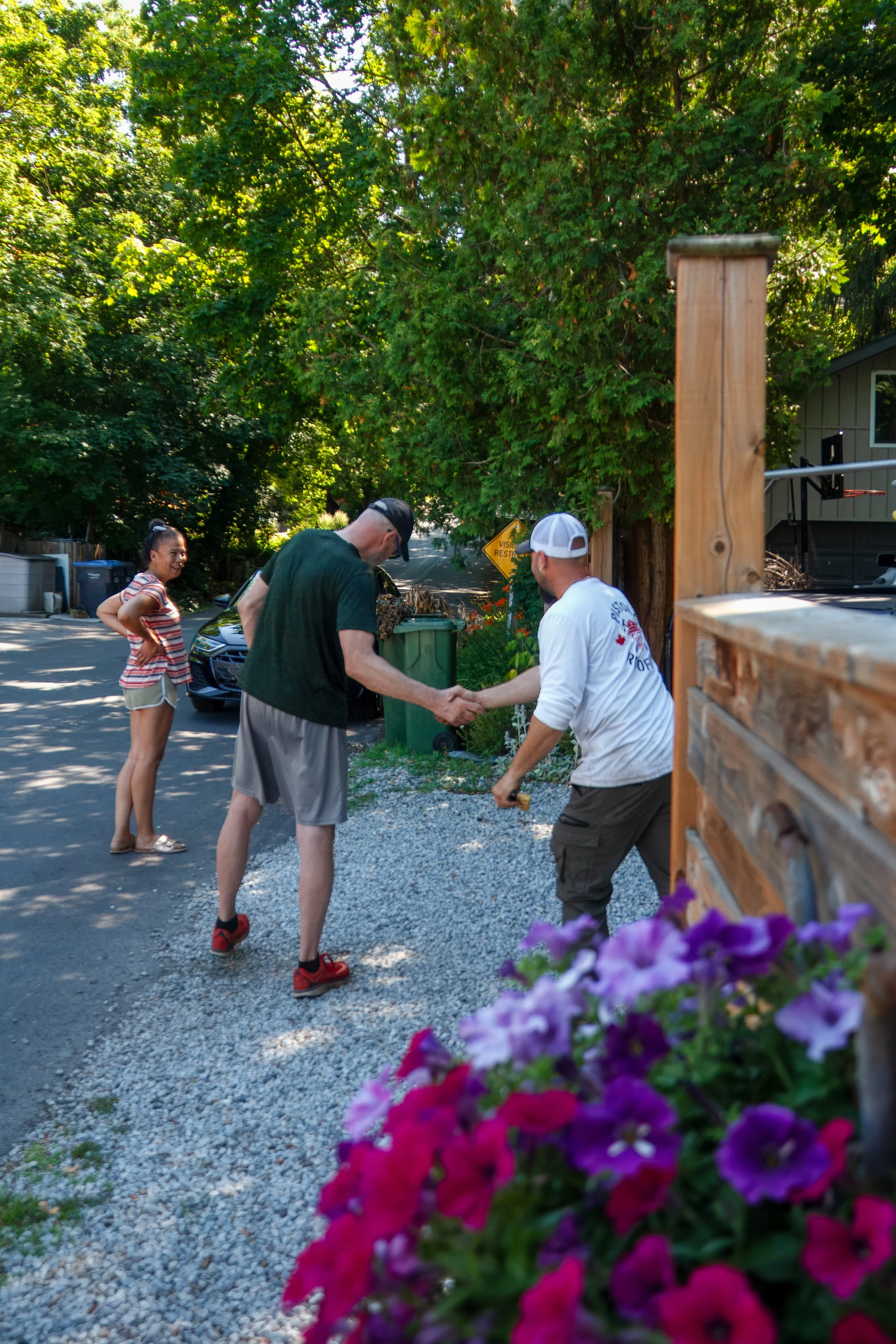 Two men shaking hands outdoors on a sunny day while a young woman watches and smiles nearby. Colorful flowers are in foreground, trees are in background.