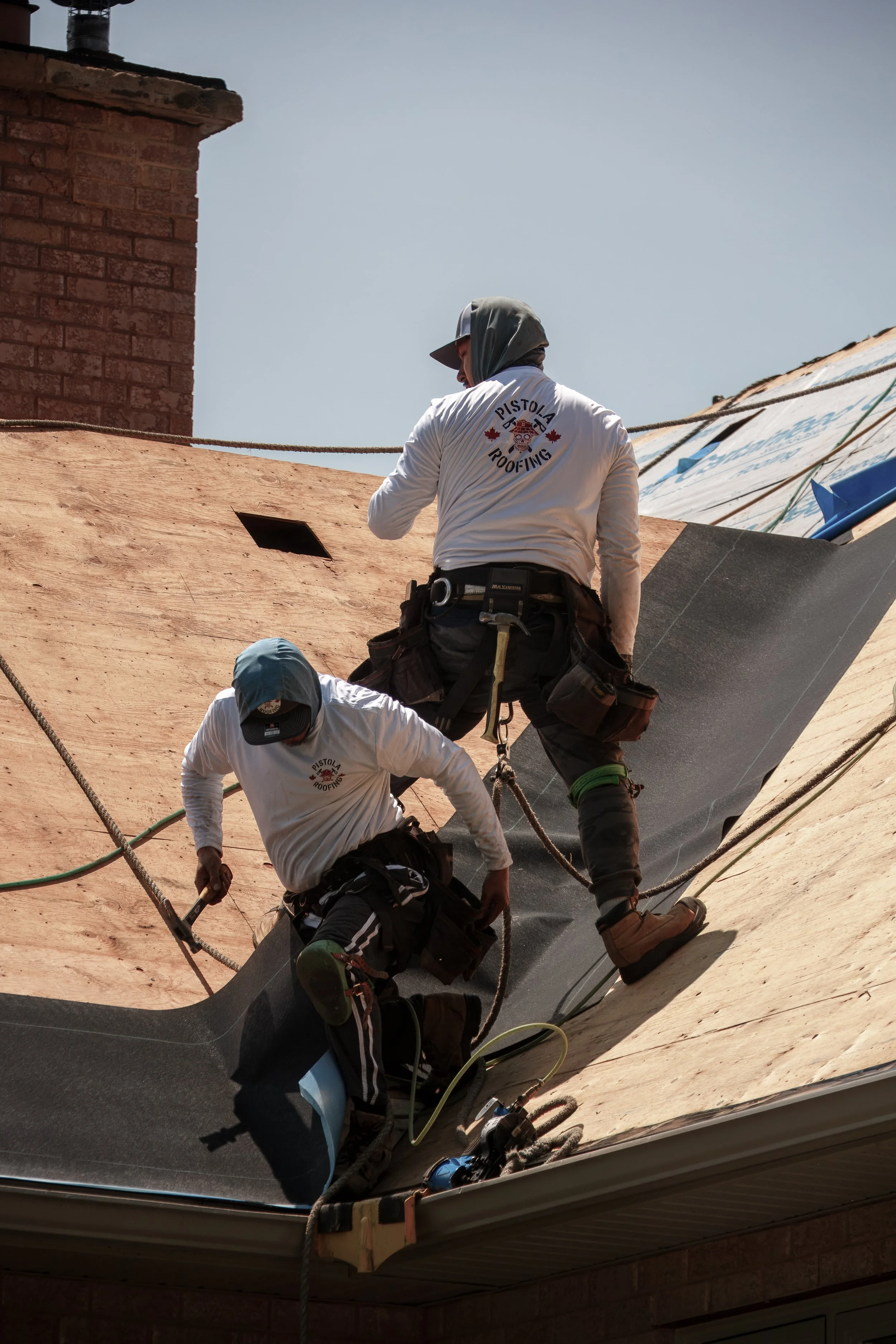 Two workers on a roof installing roofing material, wearing work gear and tools.
