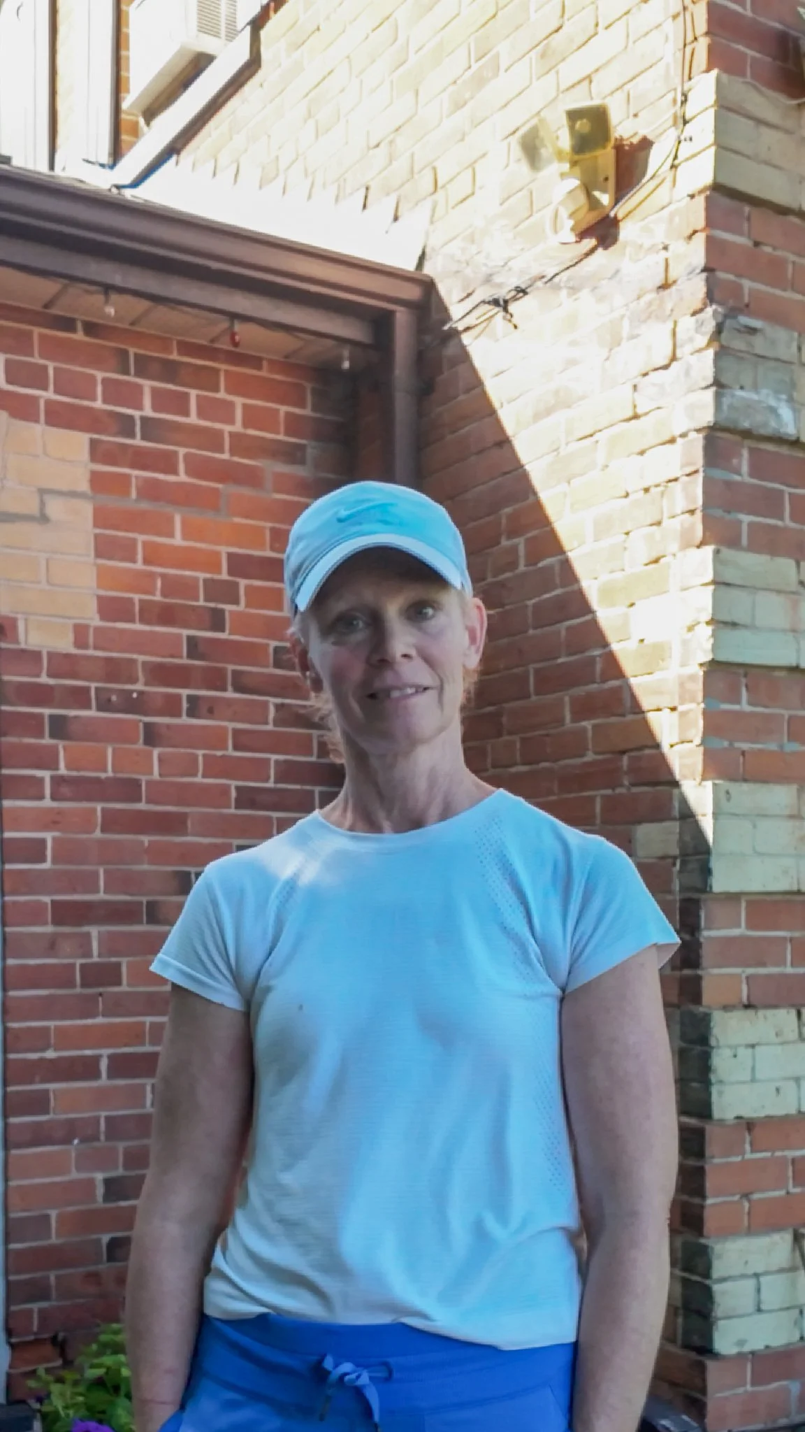 A woman wearing a light blue cap and white athletic shirt standing in front of a brick wall.