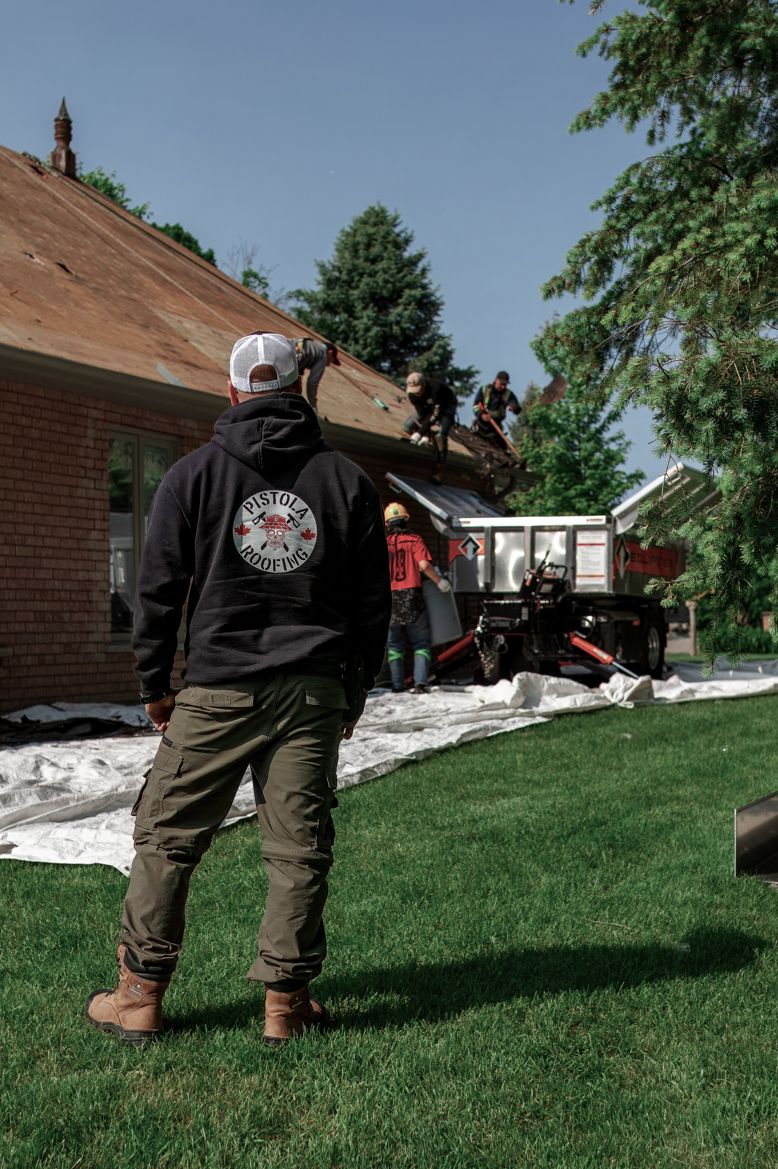 Roofers repairing a roof of a brick house while a man in a hoodie watches from the lawn.