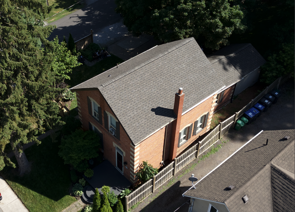 An aerial view of a two-story brick house with gray shingle roof, chimney, and a fenced backyard. Nearby are trees, a driveway with trash bins, and neighboring houses.