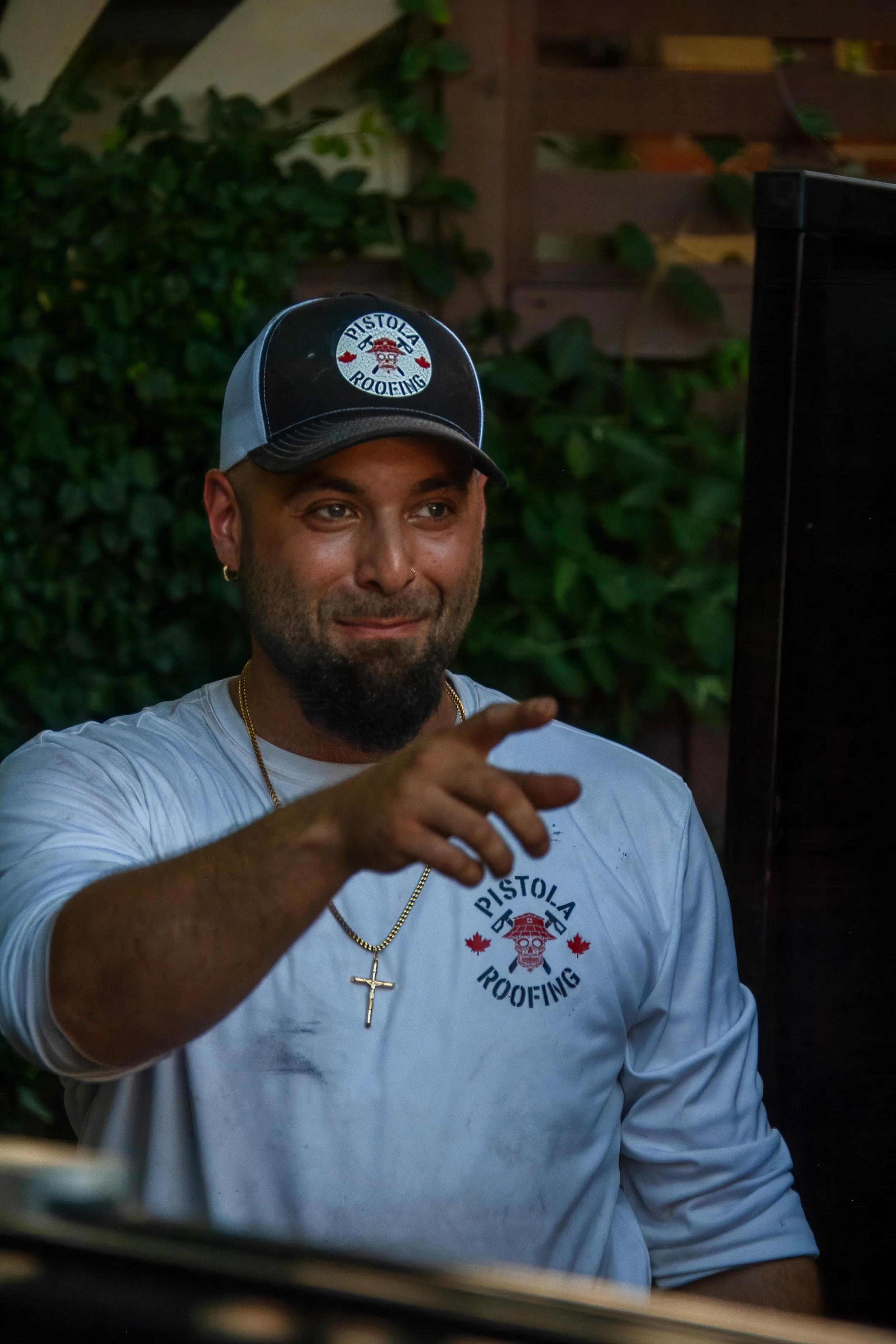 A man wearing a Pistolia Roofing hat and shirt, sitting outdoors, smiling, making a gesture with his hand.