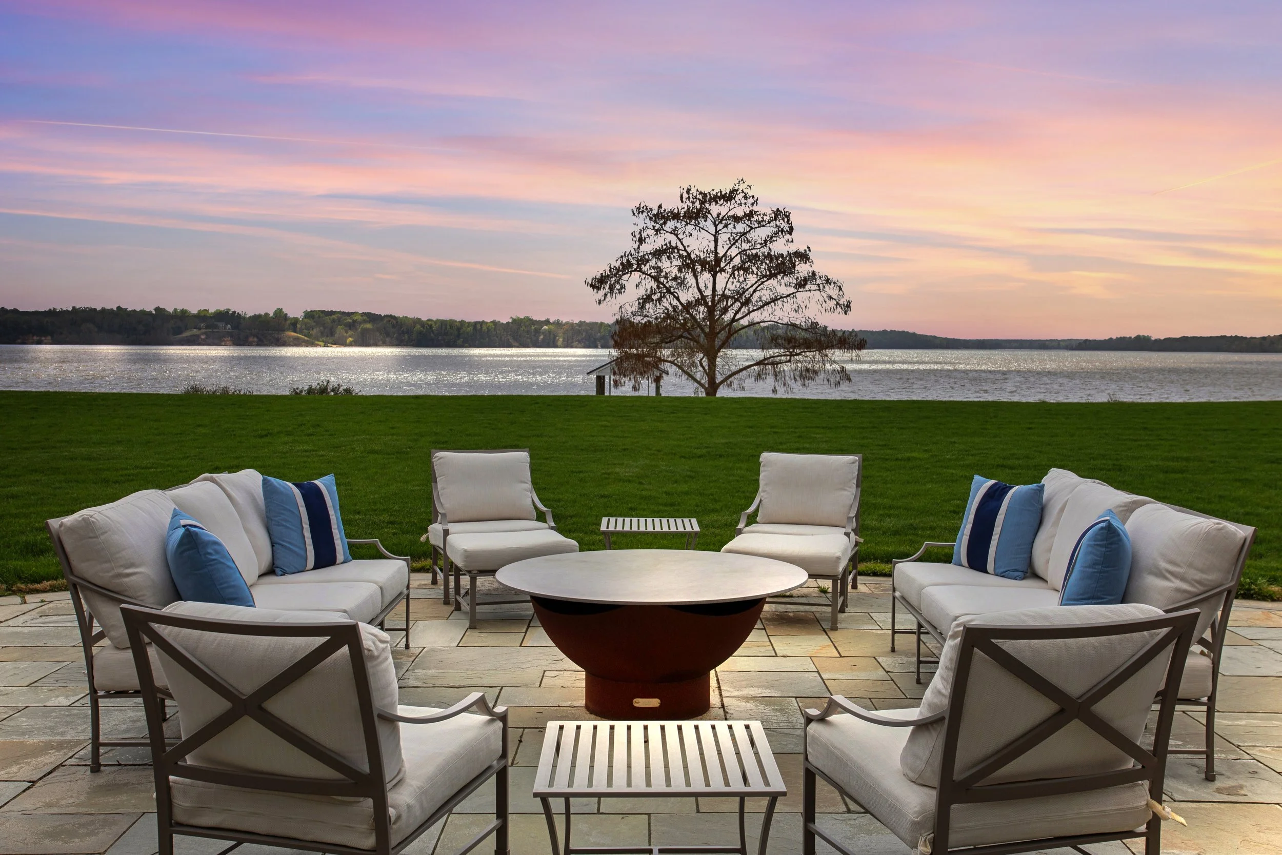 Outdoor patio seating with white cushioned chairs and sofas with blue pillows, surrounding a circular fire pit table, overlooking a grassy area and a body of water with a tree in the distance during sunset.