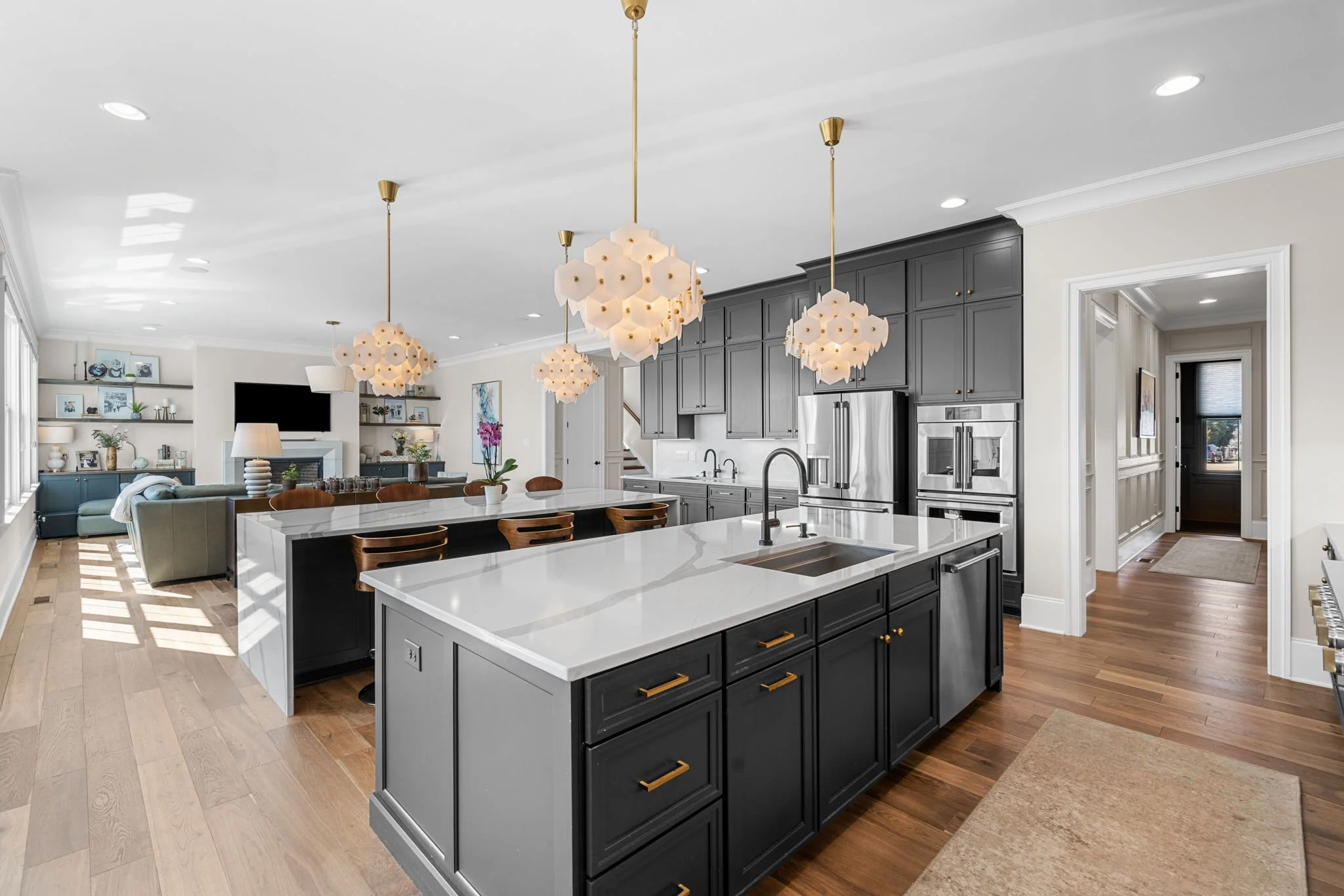 Modern kitchen with dark cabinetry, white marble countertops, and gold hardware, illuminated by decorative pendant lights, with an open living space in the background.