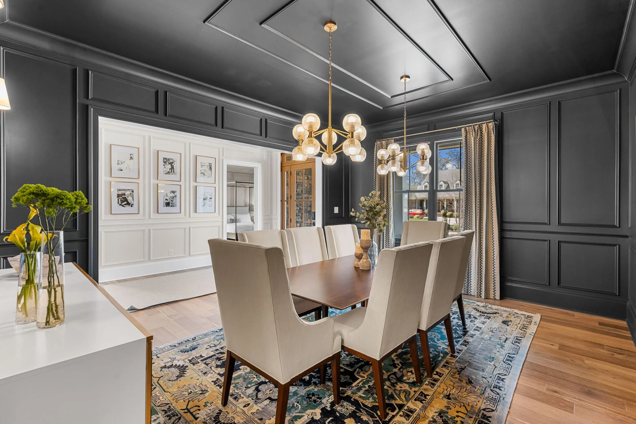 Dining room with dark gray paneled walls, six cream upholstered chairs around a wooden table, modern chandelier, large window with patterned curtains, and artwork on white wall.