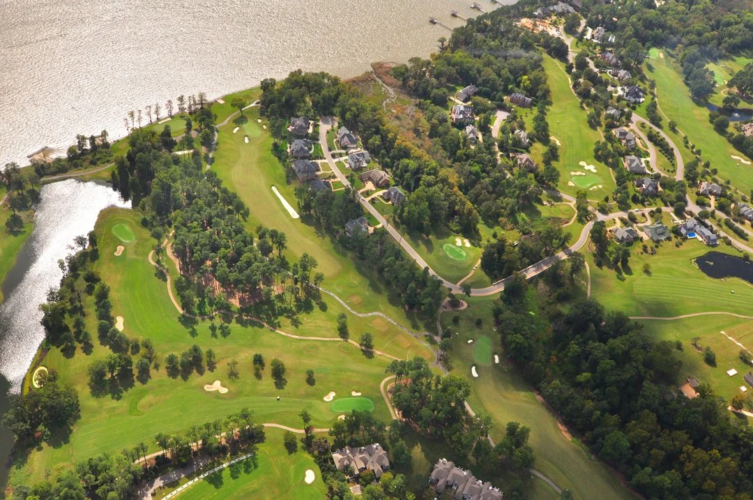 Bird's eye view of a golf course surrounded by trees, water, and residential buildings.