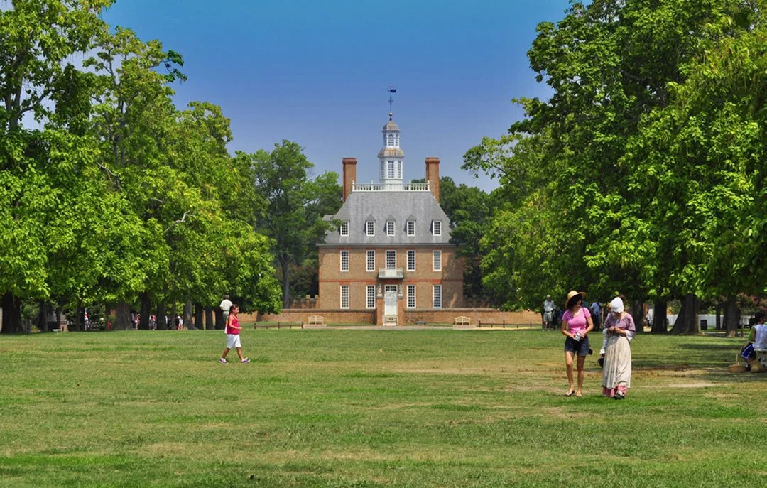 People walking on a grassy park with tall trees, overlooking a historic brick building with a courthouse steeple on a sunny day.
