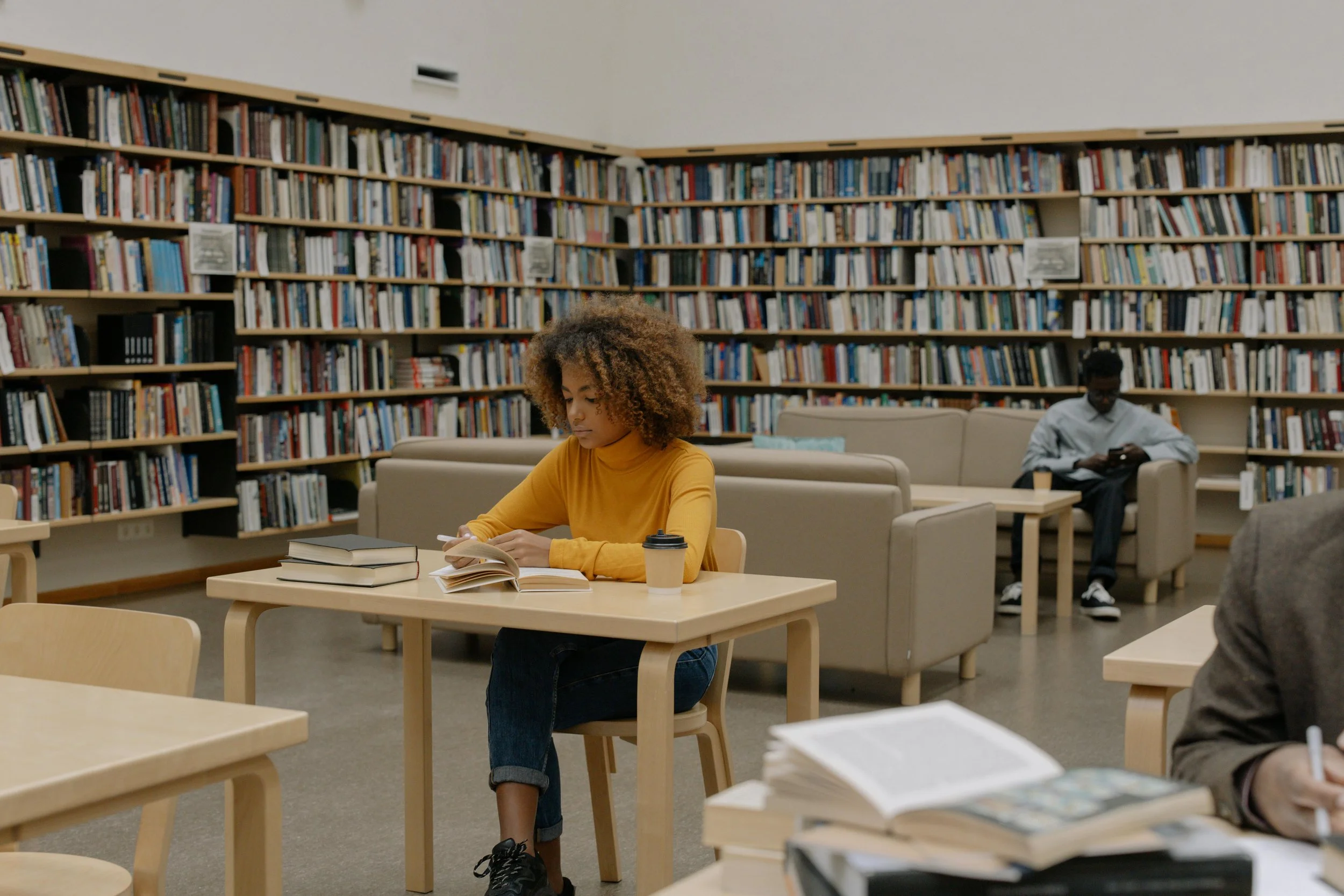 A young woman with curly hair wearing a yellow long-sleeve shirt sits at a wooden table in a library, reading a book, with a cup of coffee and stacked books nearby. In the background, a man with glasses in a light-colored shirt sits on a sofa, looking at his phone, with bookshelves filled with books behind him.