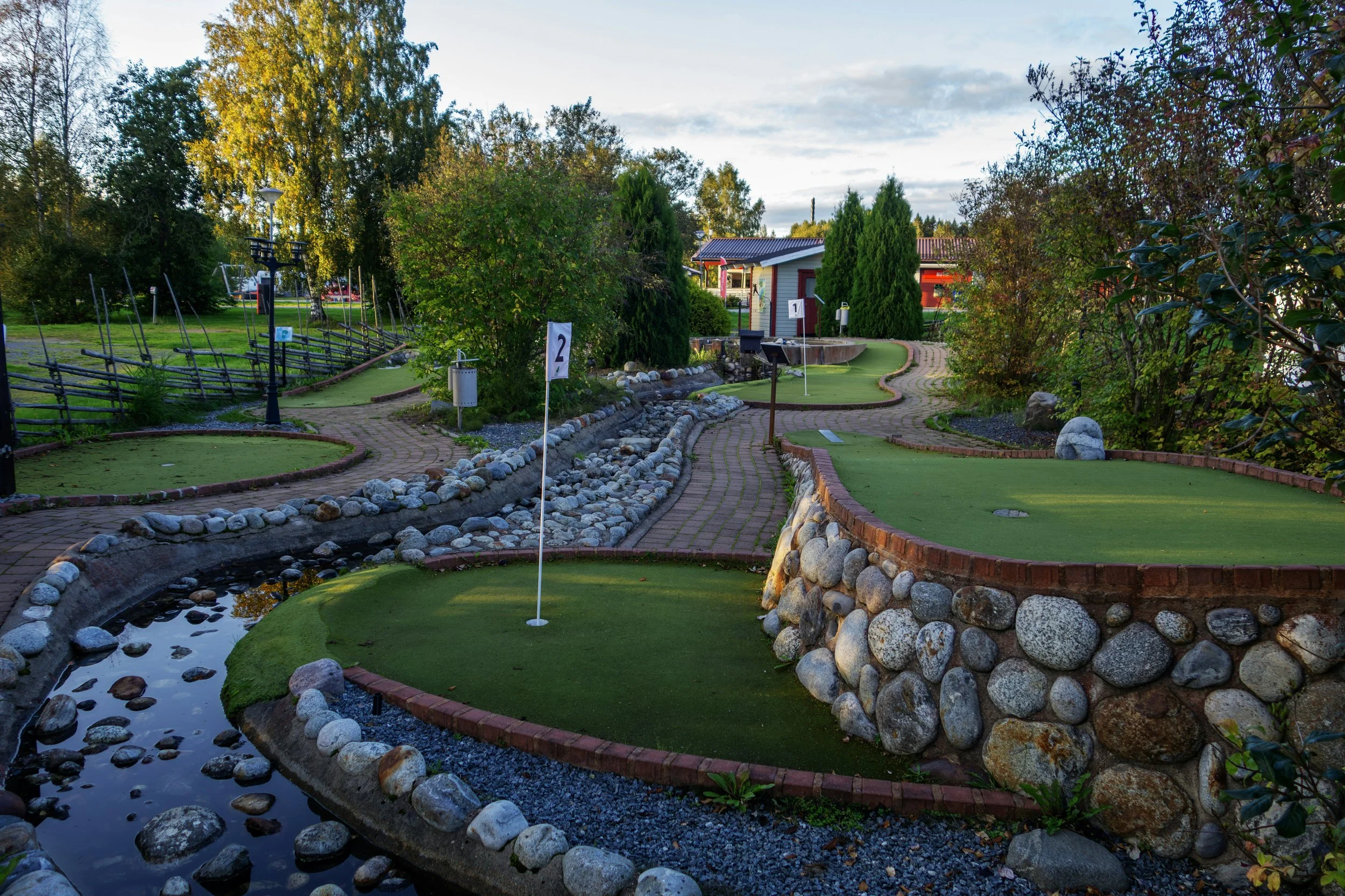 Miniature golf course with multiple holes, trees, and a small stream, under a partly cloudy sky.